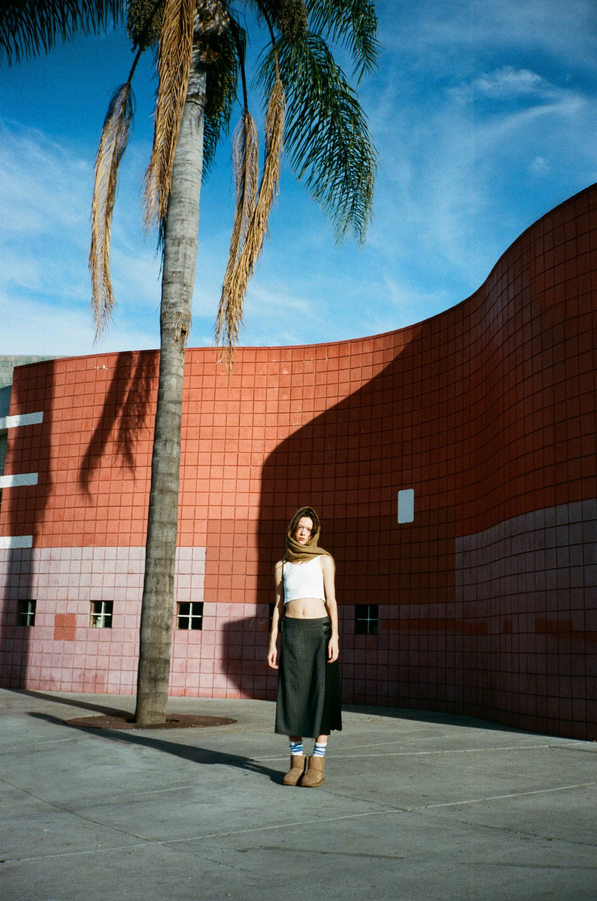 A woman wearing a white crop top, a long black skirt, striped socks, and tan shoes stands on a sidewalk in front of a pink curved building with small windows and a tall palm tree, under a blue sky with some clouds.