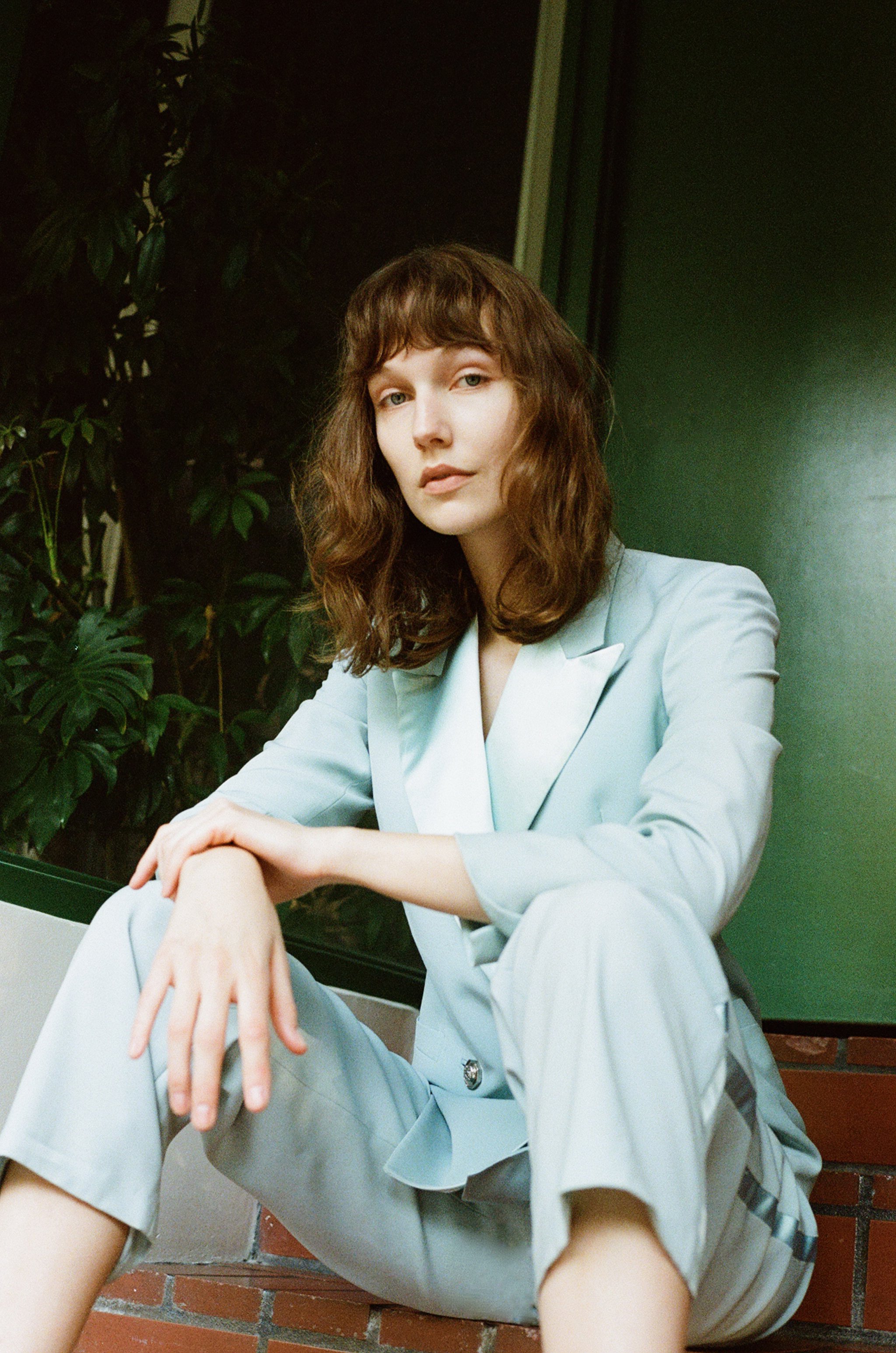 Young woman with wavy brown hair, sitting on a brick step, wearing a light gray suit, with a green wall and plant in the background.