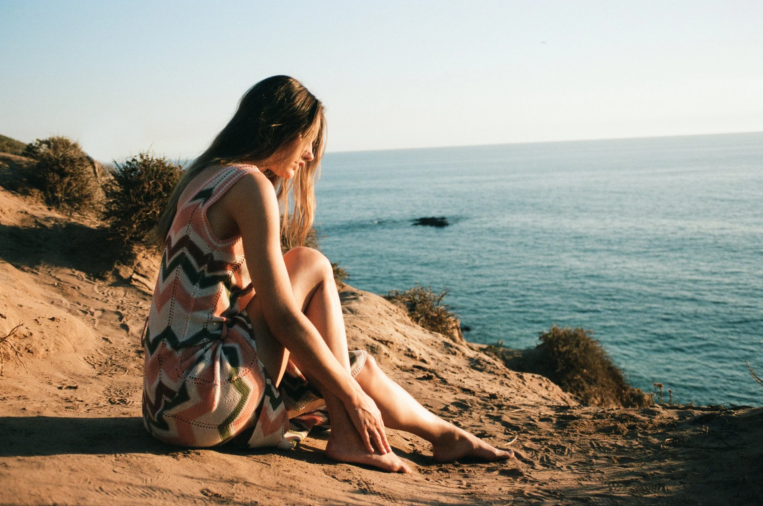 A woman sitting on a sandy cliff by the ocean, looking down, wearing a colorful patterned dress, with the sea and sky in the background.