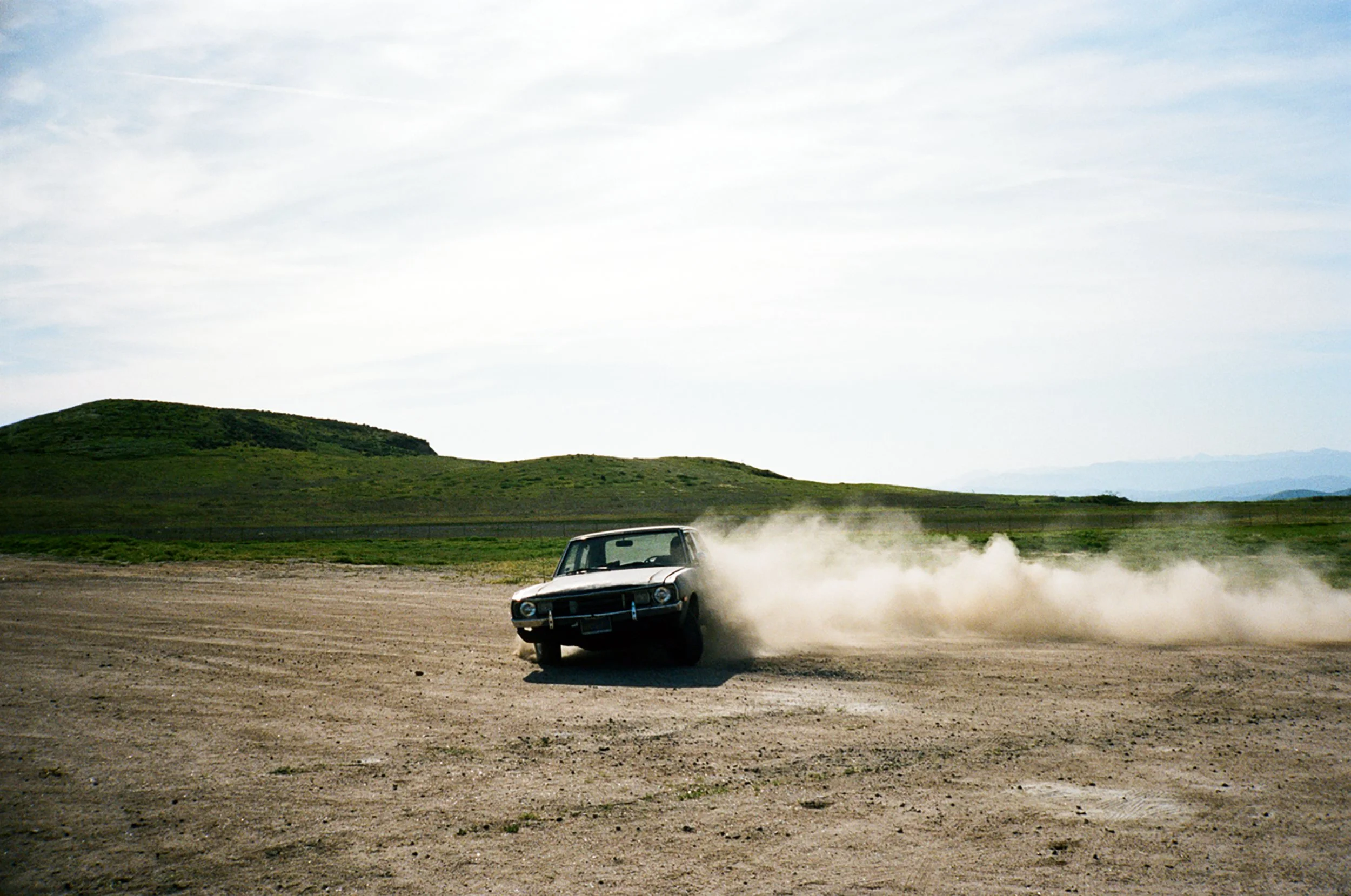 A car kicking up dust as it drifts on a dirt field with green hills and a cloudy sky in the background.