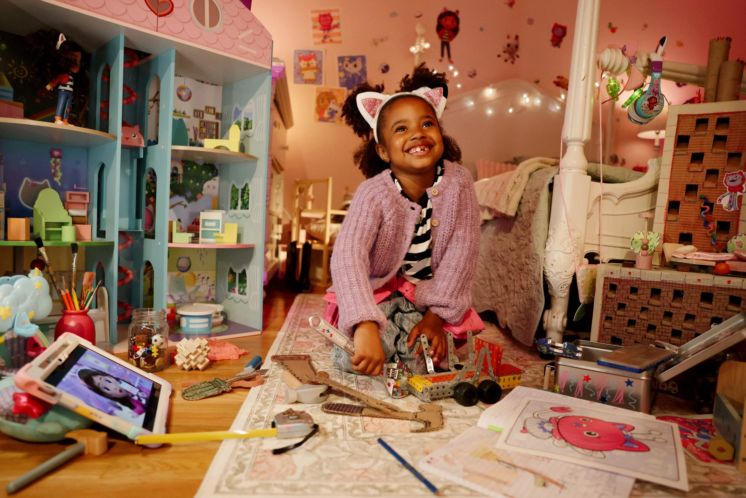 A young girl with curly hair, wearing cat ear headbands, purple cardigan, and pink skirt, smiling while sitting on the floor amidst toys, craft supplies, and coloring pages in a colorful children's playroom decorated with cartoon character posters on