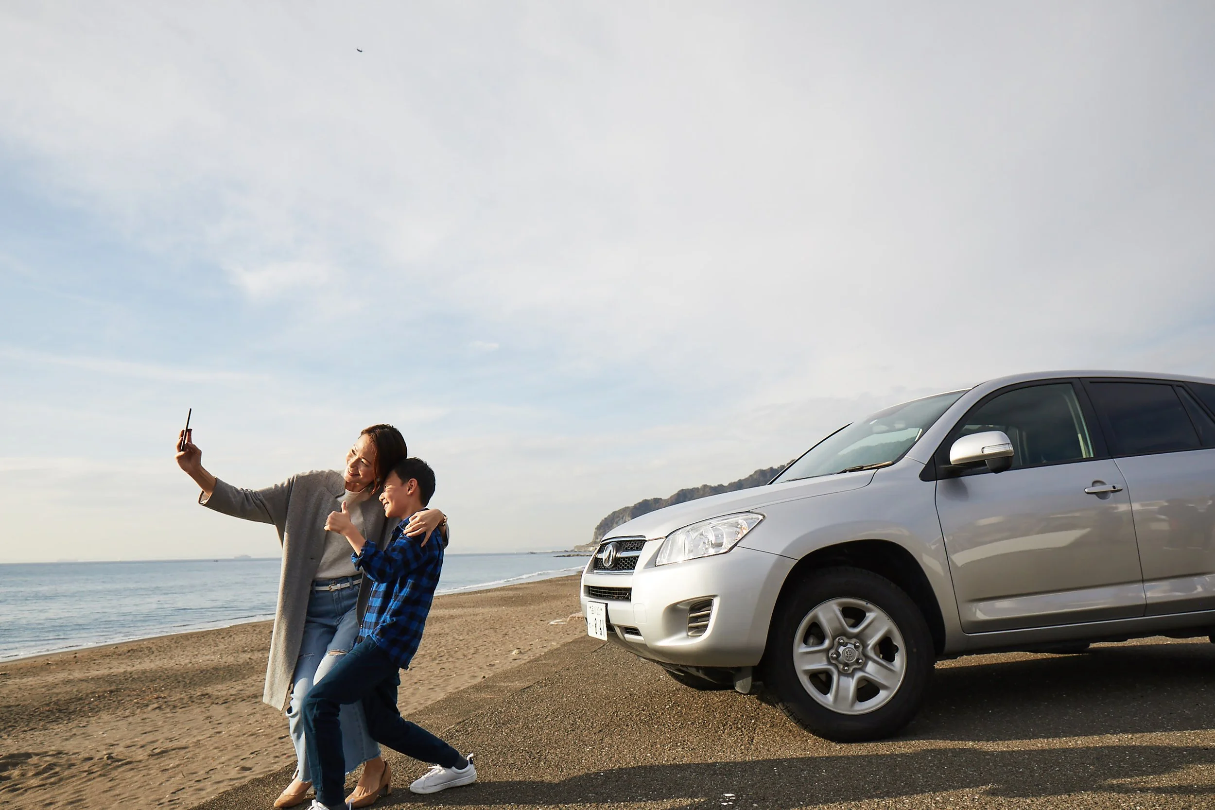 A woman and a boy taking a selfie on the beach with a gray car parked nearby, the ocean and sky in the background.