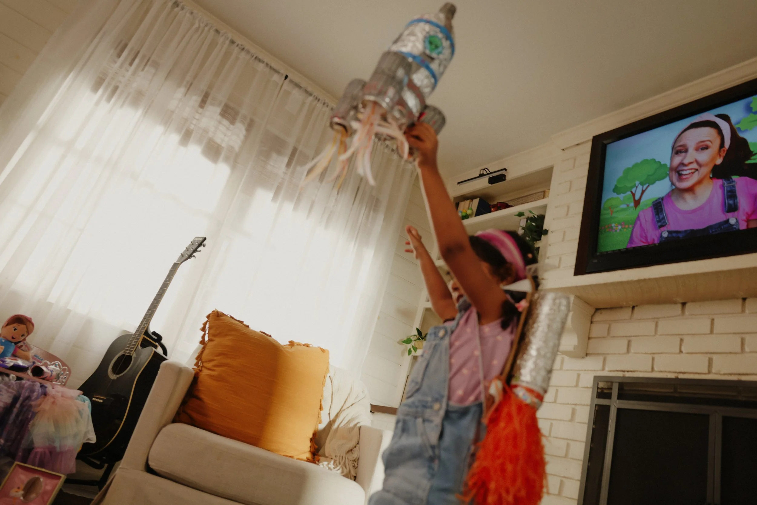 Child raising a colorful spaceship craft made of toilet paper rolls in a living room with a guitar, TV, and doll collection.