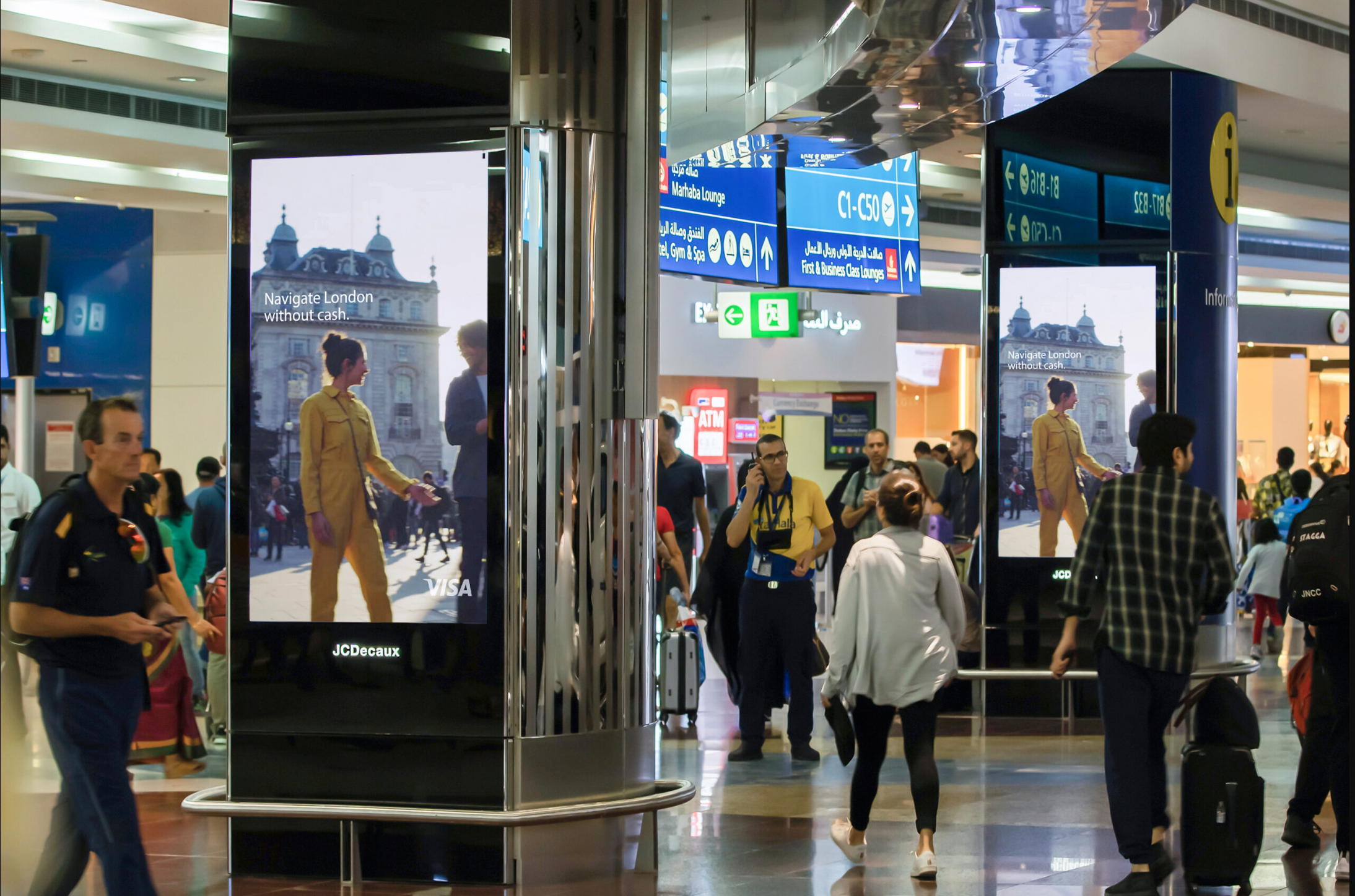 Crowd of travelers walking through an airport with digital signage and directional signs overhead.