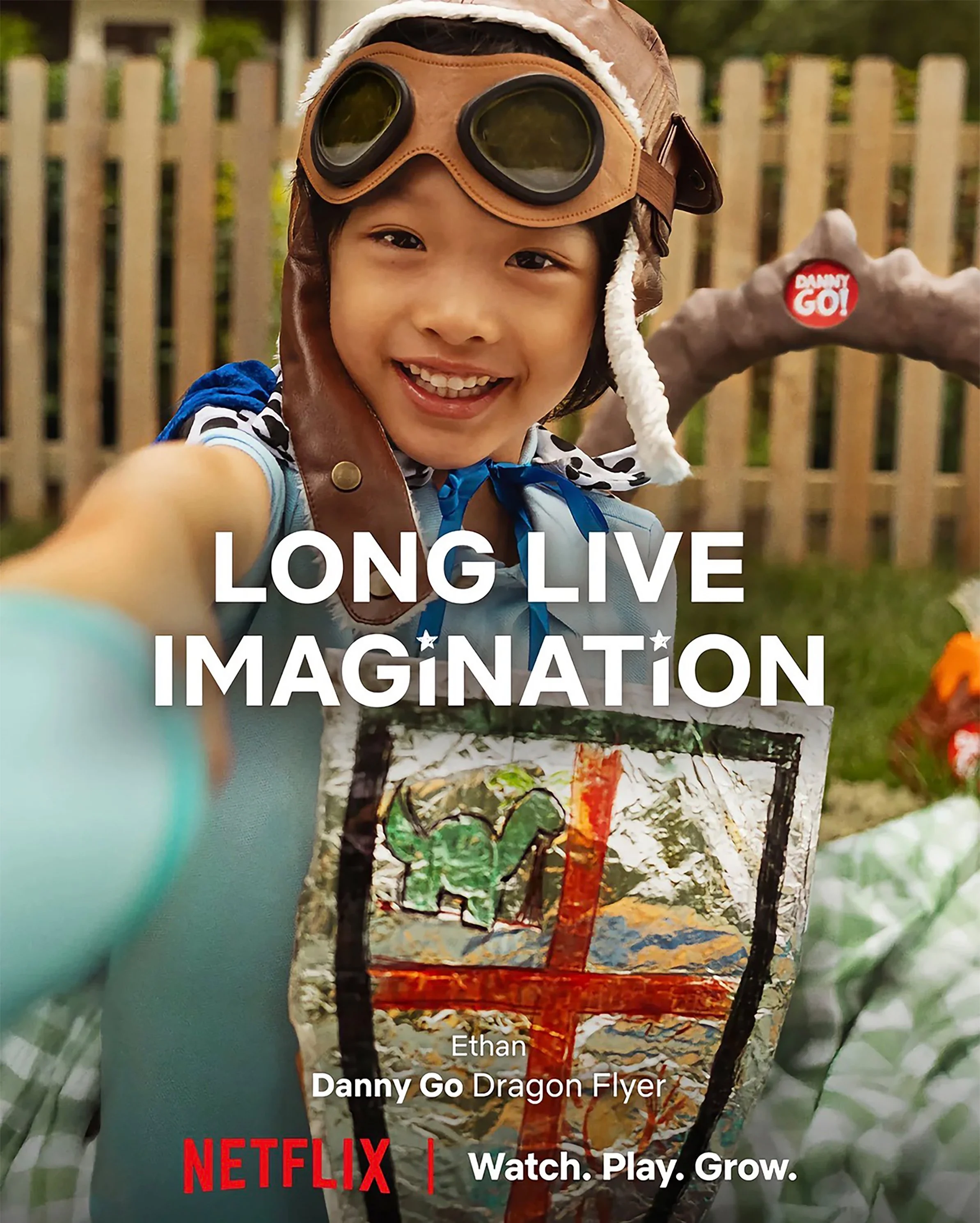 A young boy wearing a brown aviator hat and goggles smiling and taking a selfie outdoors. The background features a wooden fence and green foliage, with text overlay promoting Netflix and encouraging viewers to watch, play, and grow.