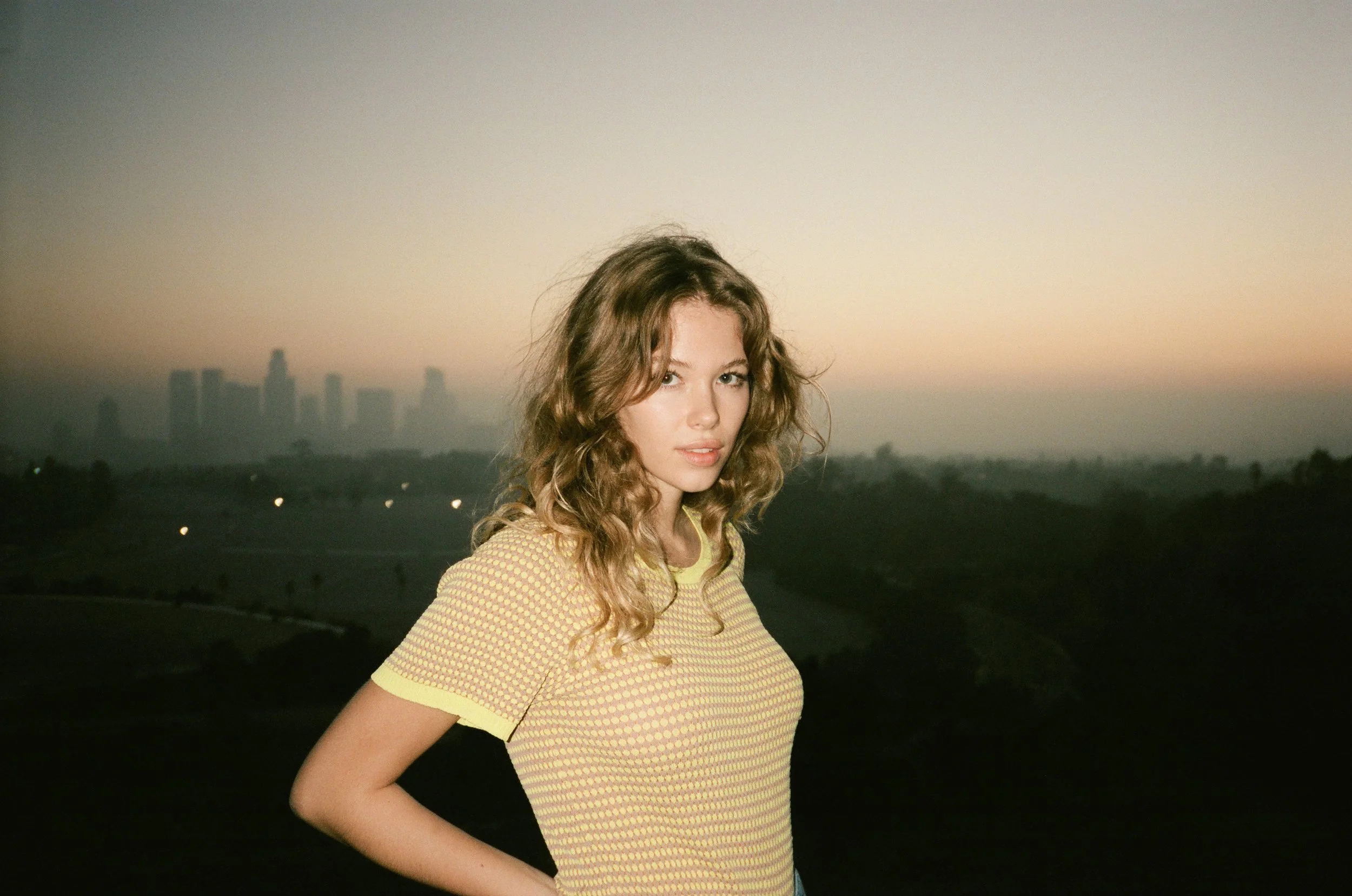 Young woman with wavy blond hair, wearing a yellow patterned shirt, standing outdoors at dusk with a city skyline in the background.