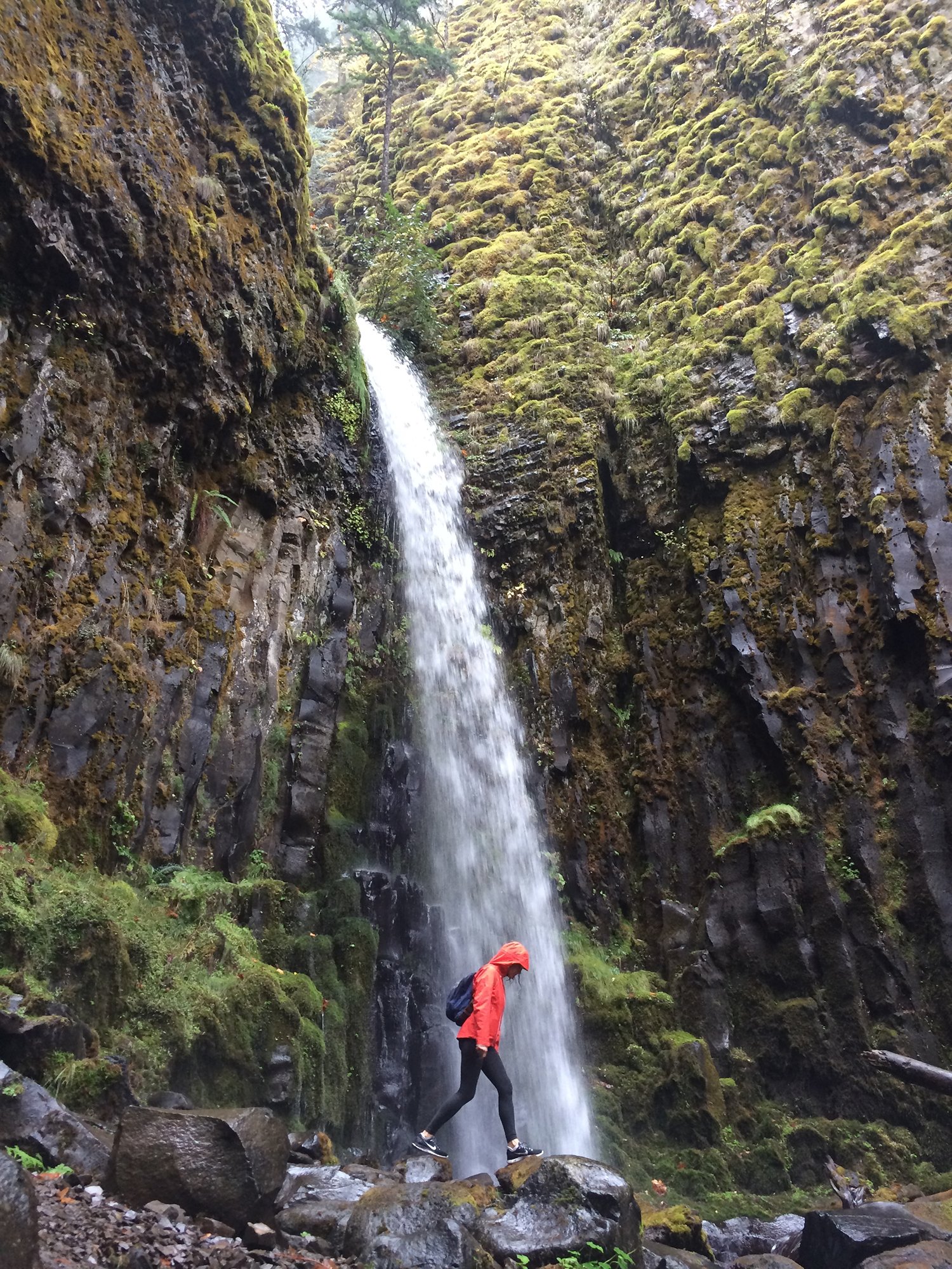 Person in red rain jacket and black leggings crossing wet rocks near a waterfall in a mossy forest.