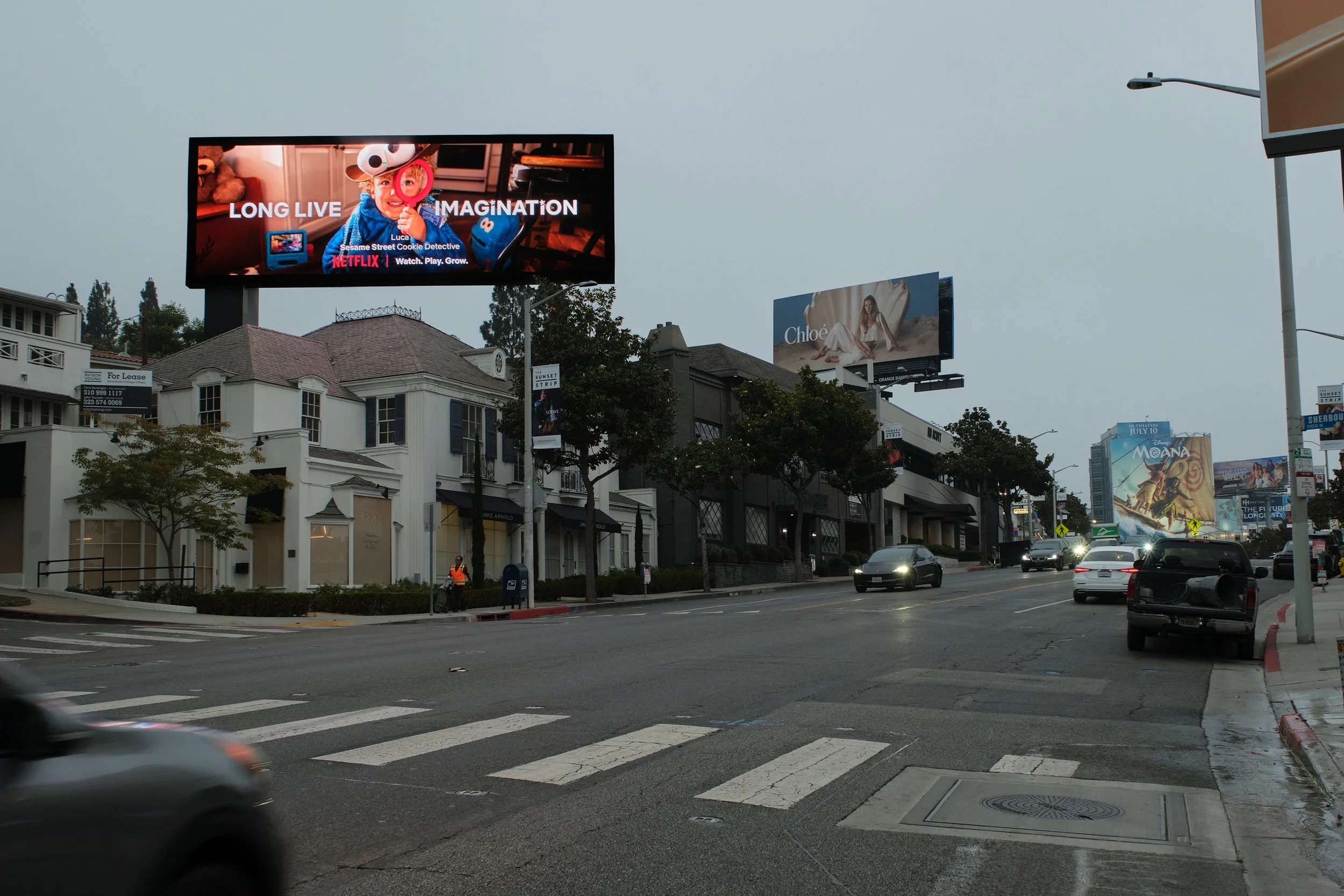 City street scene with electronic billboards, cars, trees, and sidewalks on an overcast day.