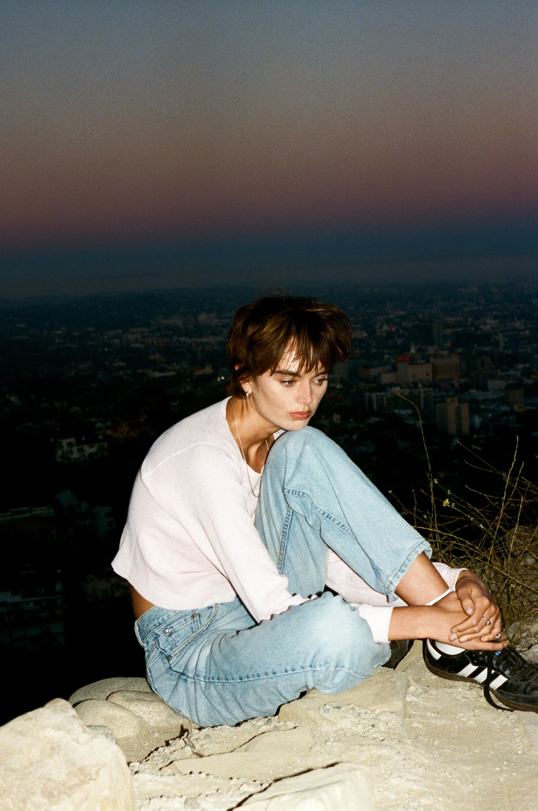 A woman with short brown hair sitting on a rocky surface during dusk, with a cityscape in the background and a gradient sky from pink to dark blue.