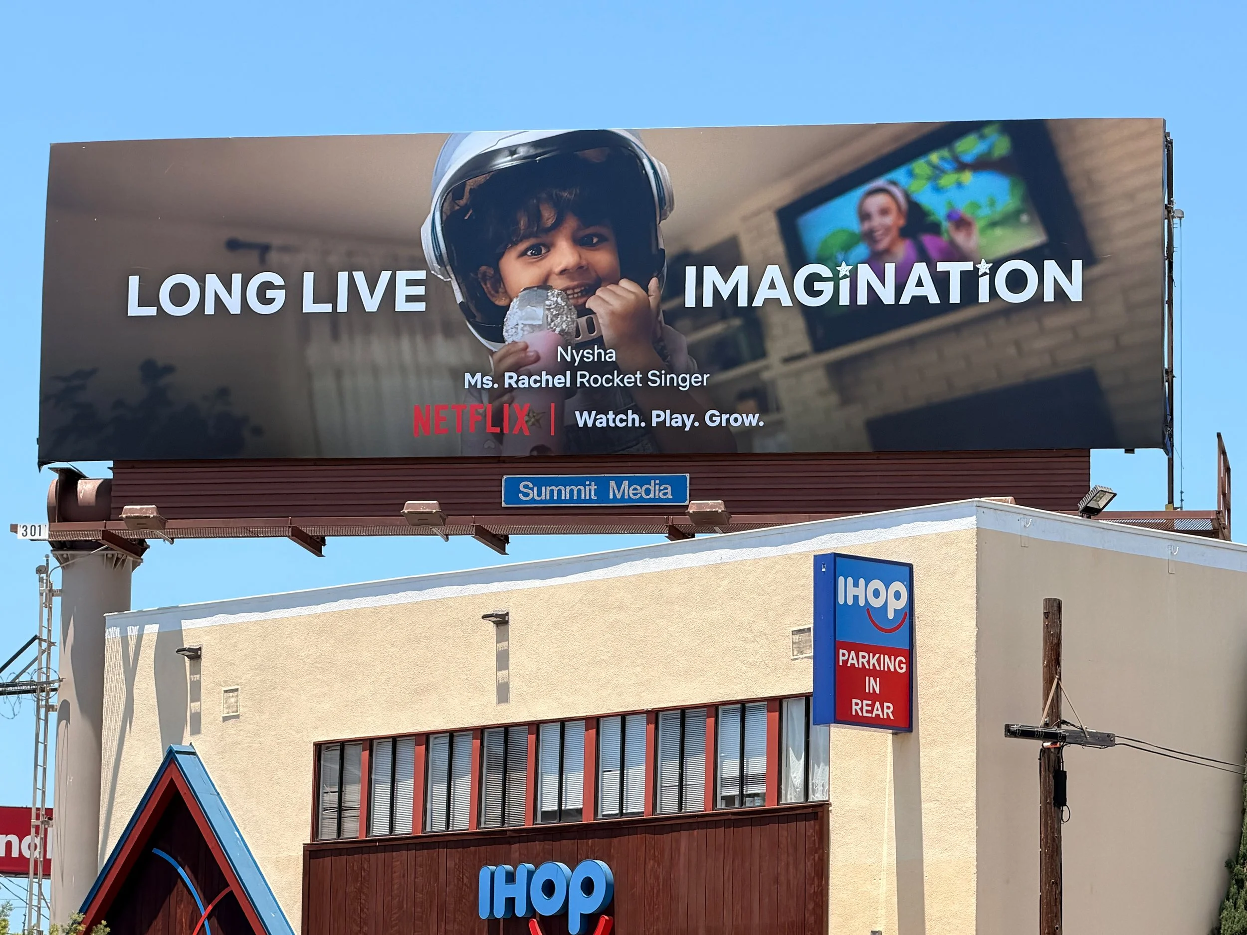 Billboard featuring a young boy with curly hair wearing a helmet and holding a rock, with the words "LONG LIVE IMAGINATION" and a Netflix logo, alongside an image of a girl displayed on a screen in the background.