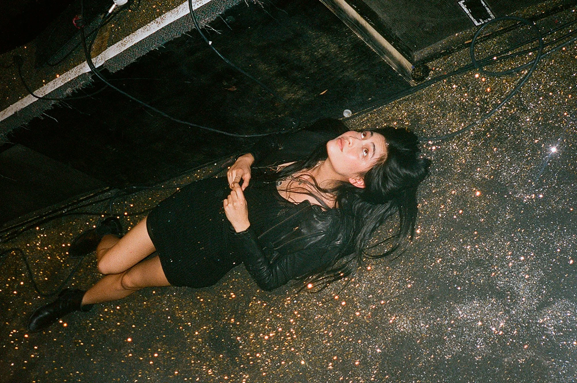 A woman with long black hair lying on a floor covered in gold glitter, looking upward, with black clothing and a black leather jacket, surrounded by cables and equipment.