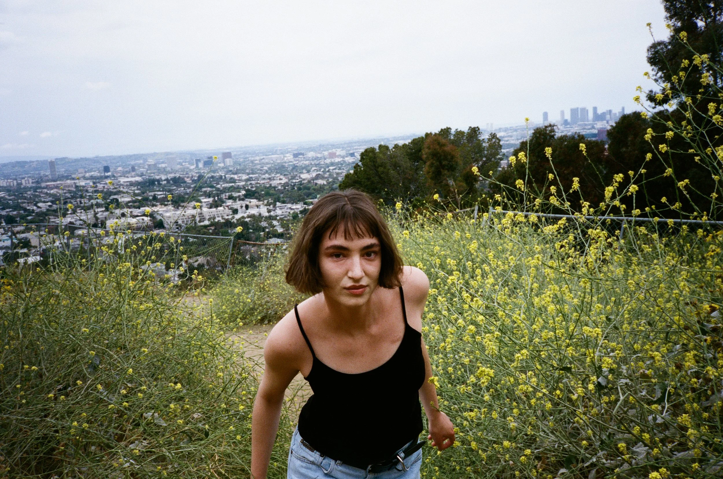 Young woman with short brown hair in a black tank top and jeans walking through yellow wildflowers on a hillside with a cityscape in the background.