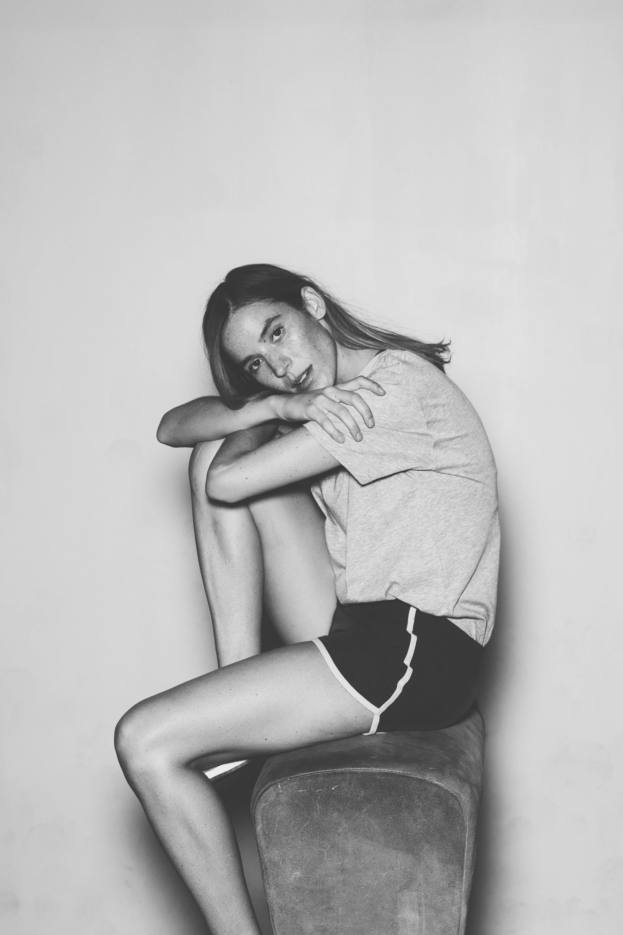 A young woman with freckles and dark hair, sitting on a cushioned stool against a plain wall, wearing a t-shirt and shorts, with her arms wrapped around her knees.