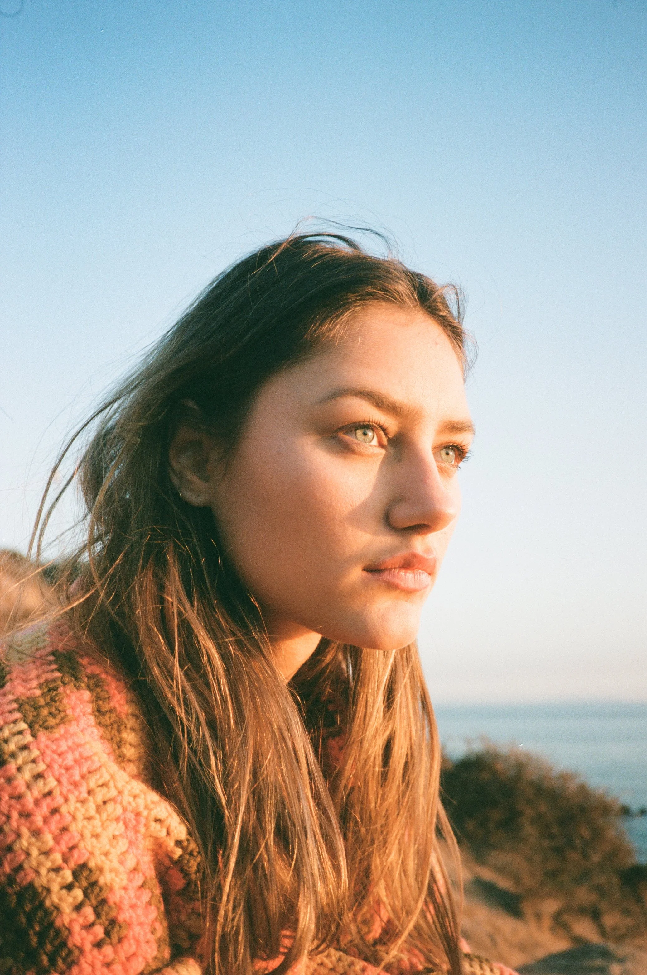 A young woman with long, brown hair and blue eyes gazing into the distance during sunset near a body of water.