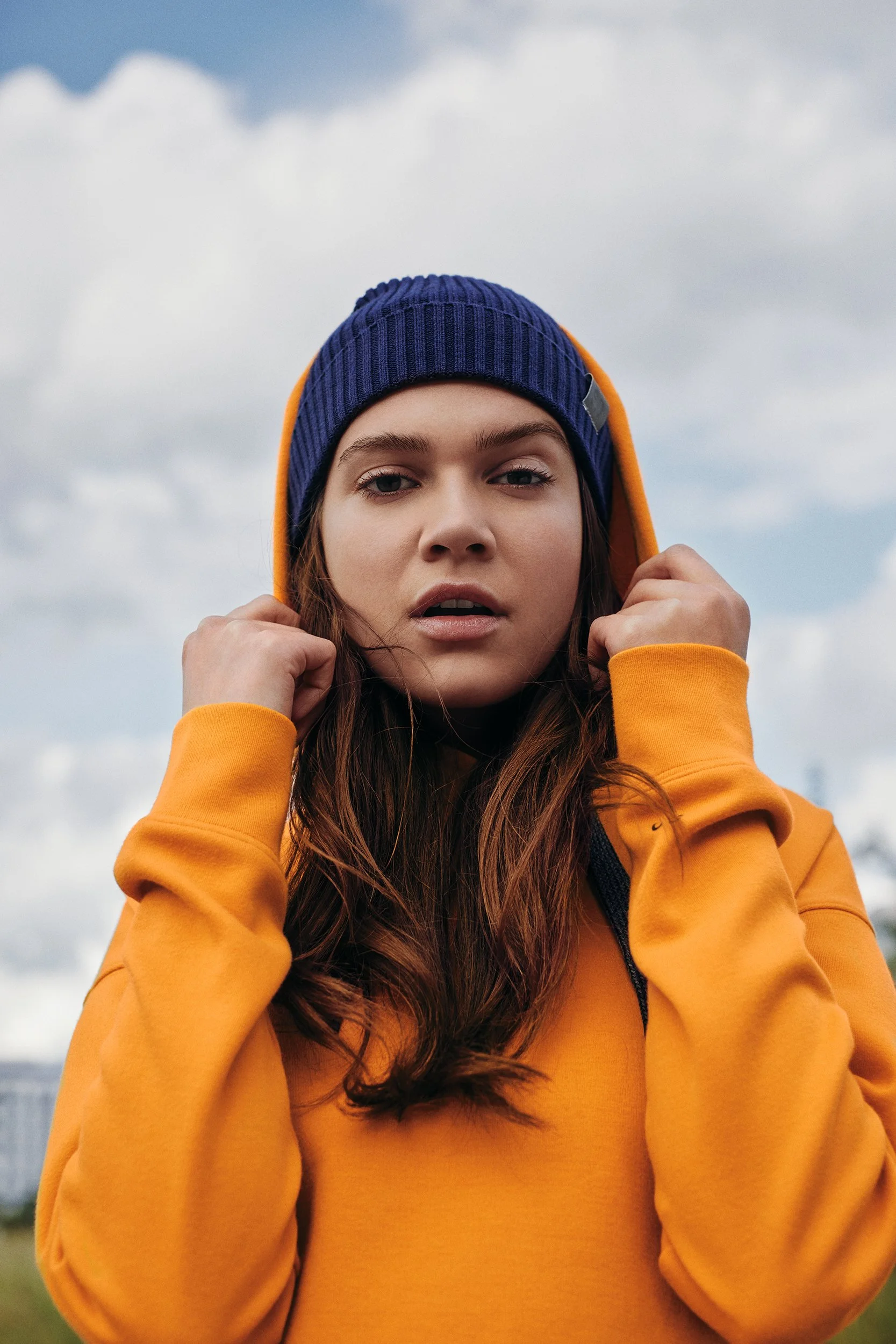 A young woman with long brown hair wearing a yellow hoodie and a blue knit beanie, standing outdoors under a cloudy sky.