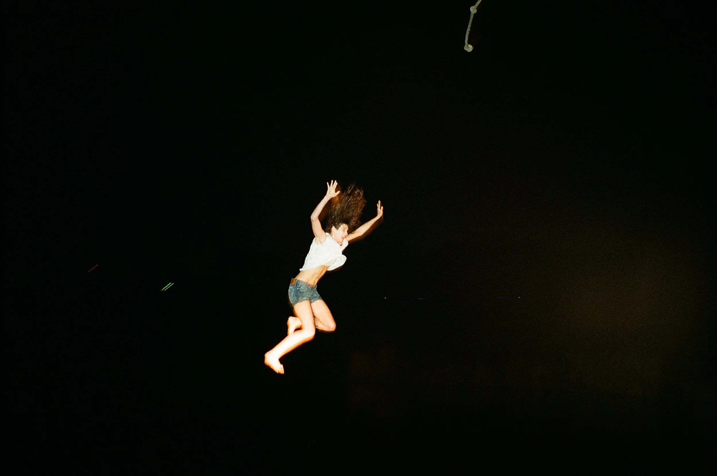 A woman falling through the night sky at night, with her arms and legs extended, wearing a white tank top and denim shorts.