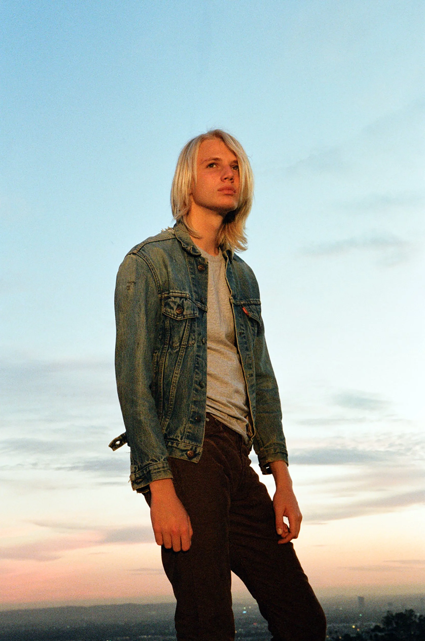 Young man with long blonde hair standing outdoors during sunset, wearing a denim jacket and dark pants.