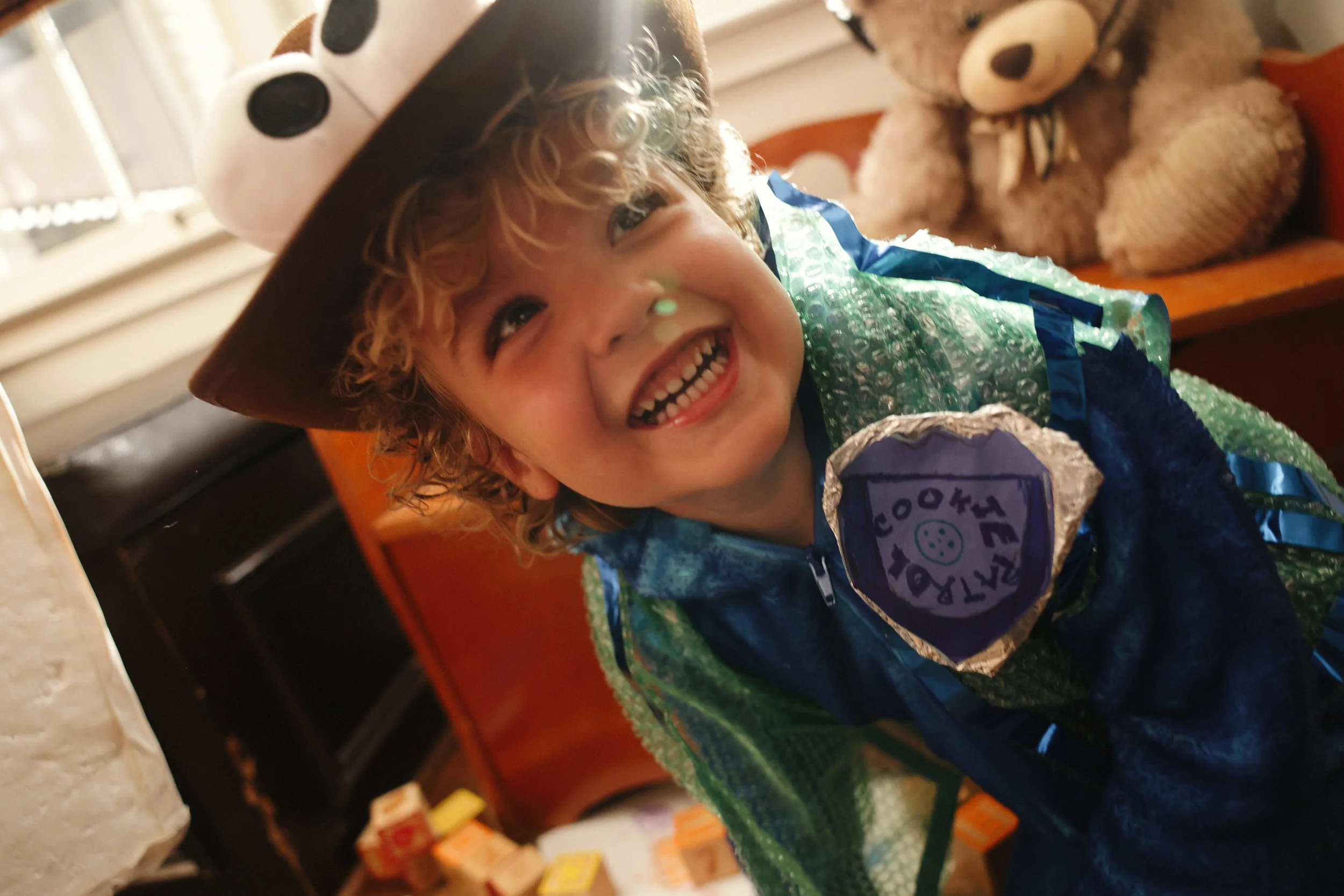 A smiling child with curly hair wearing a cowboy hat and a shiny blue and green costume, sitting in a room with a bear plush toy and wooden furniture in the background.