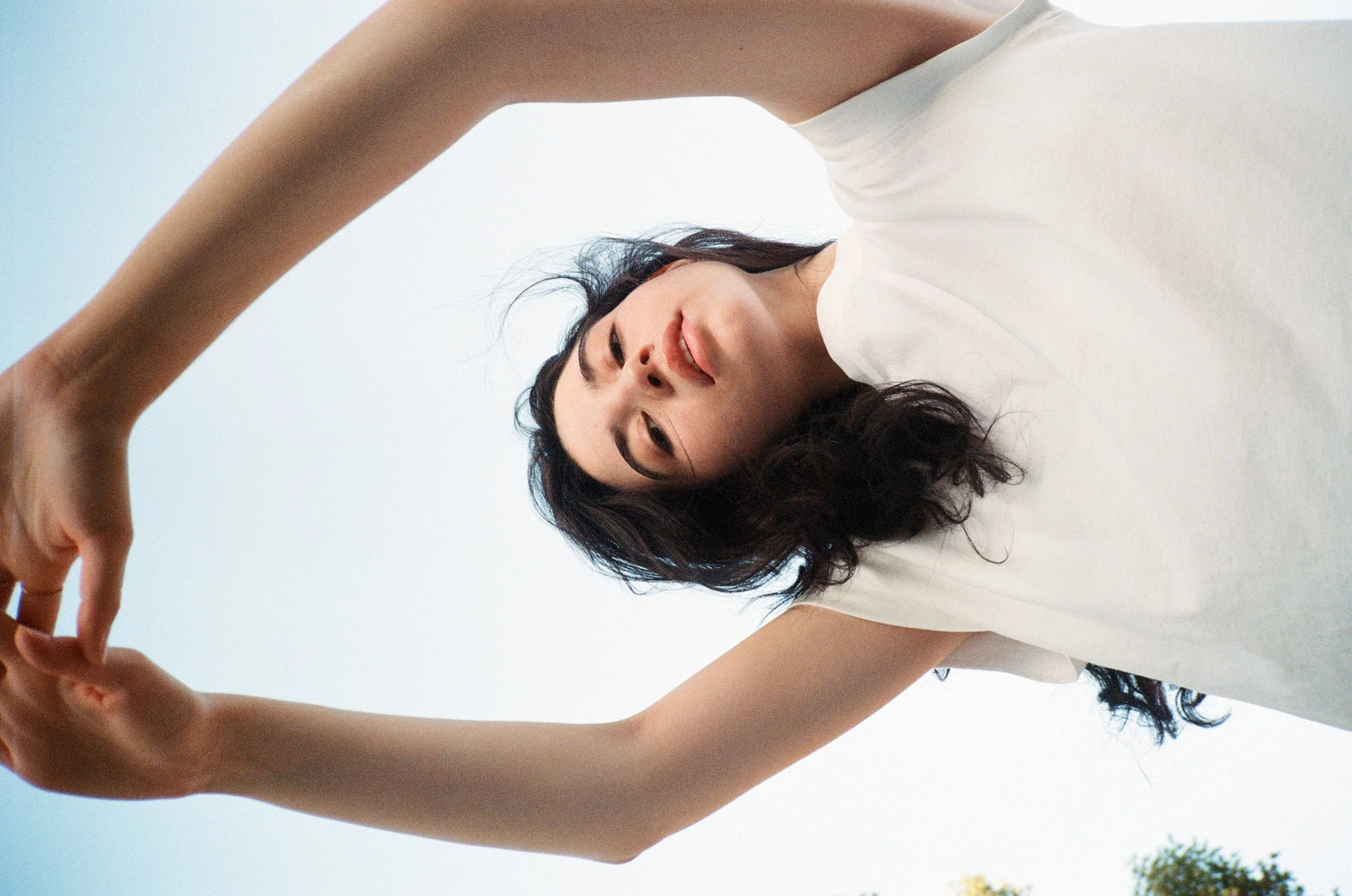 A woman with dark, wavy hair wearing a white sleeveless top reaching out and holding someone's hand, with a clear blue sky in the background.
