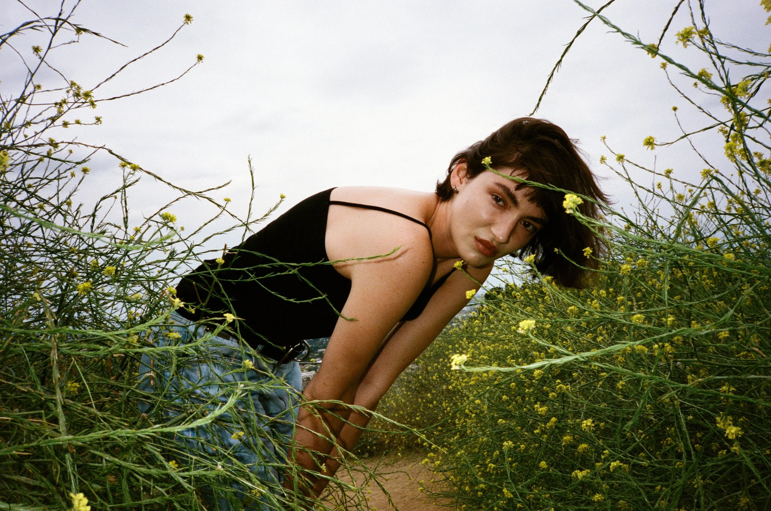 A young woman with short dark hair and a black spaghetti strap top crouches among yellow wildflowers in a field, looking at the camera with a neutral expression.