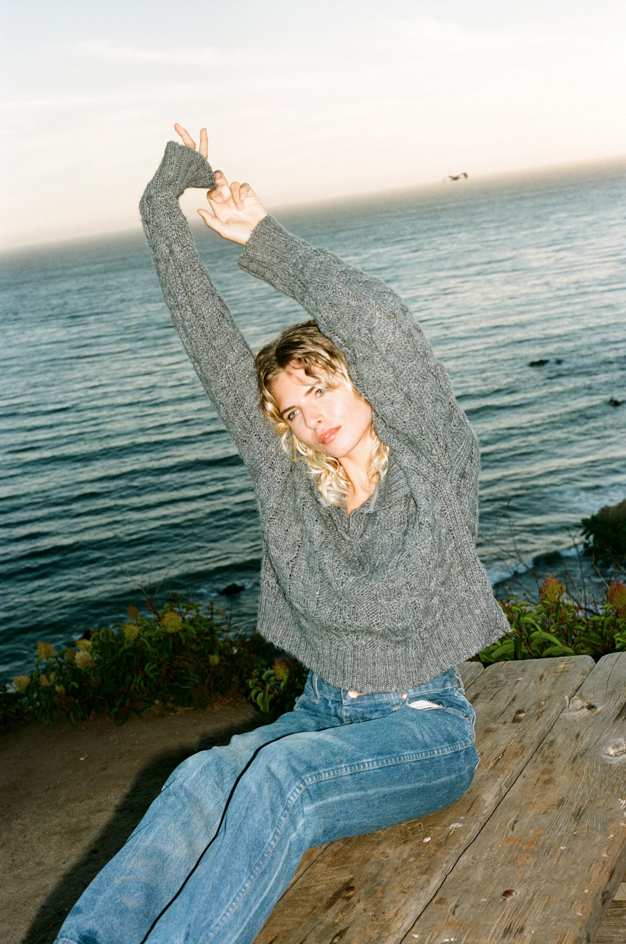 A woman in a gray sweater and jeans sitting on a wooden bench by the ocean with her arms raised above her head, stretching.