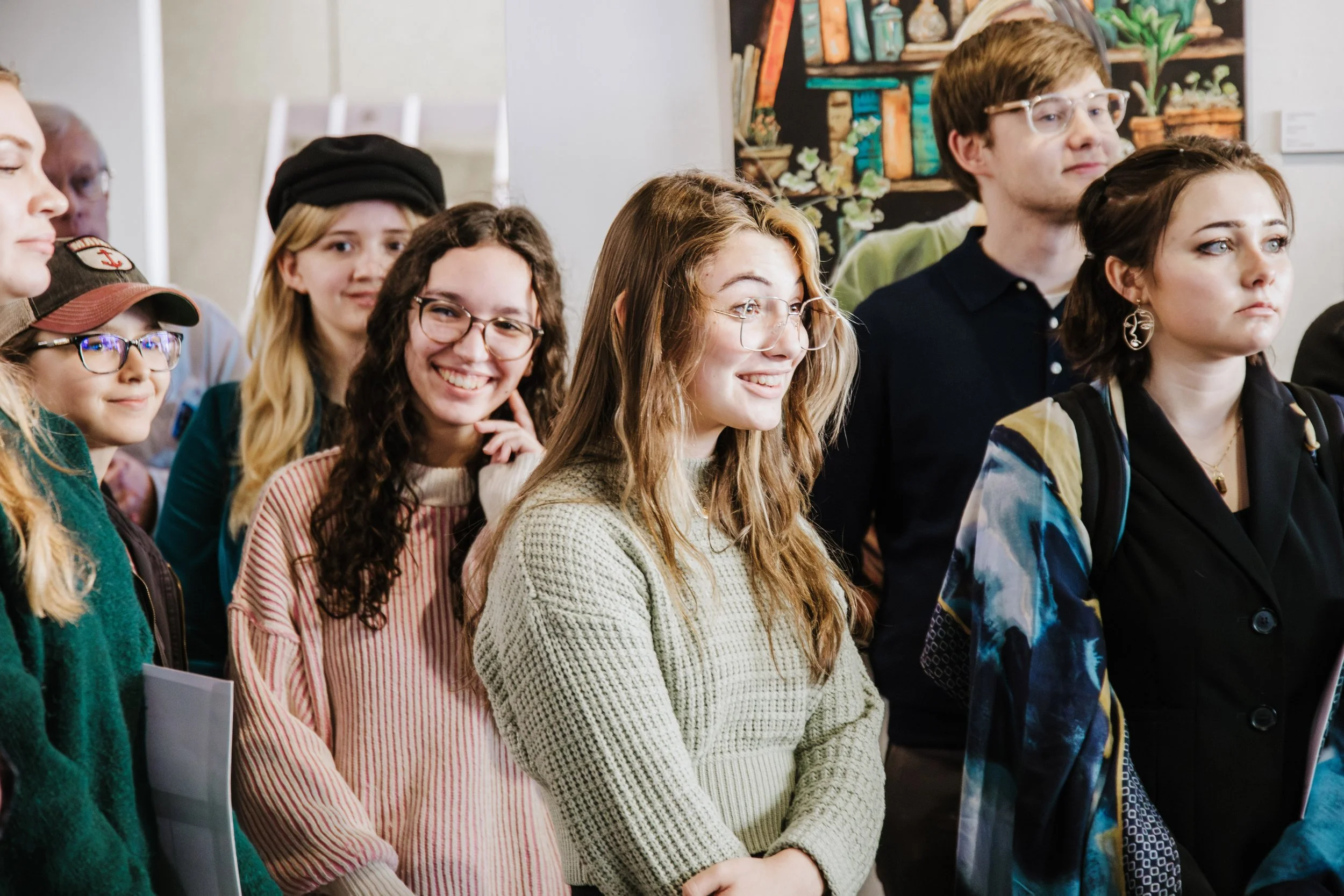 Group of young people standing together, some smiling and others observing, in an indoor setting with colorful artwork on the wall behind them.