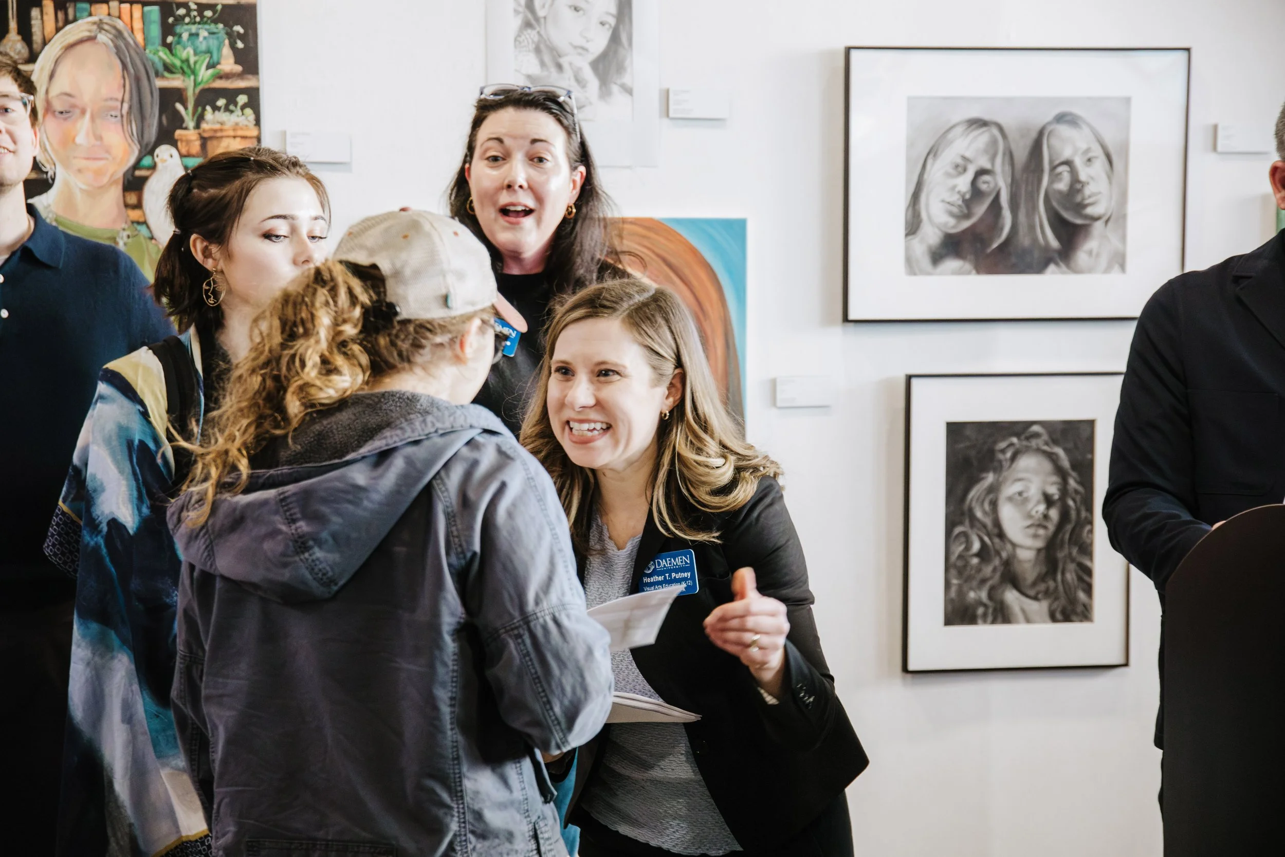 Group of people at an art gallery, engaging in conversation, with framed artwork on the wall behind them.