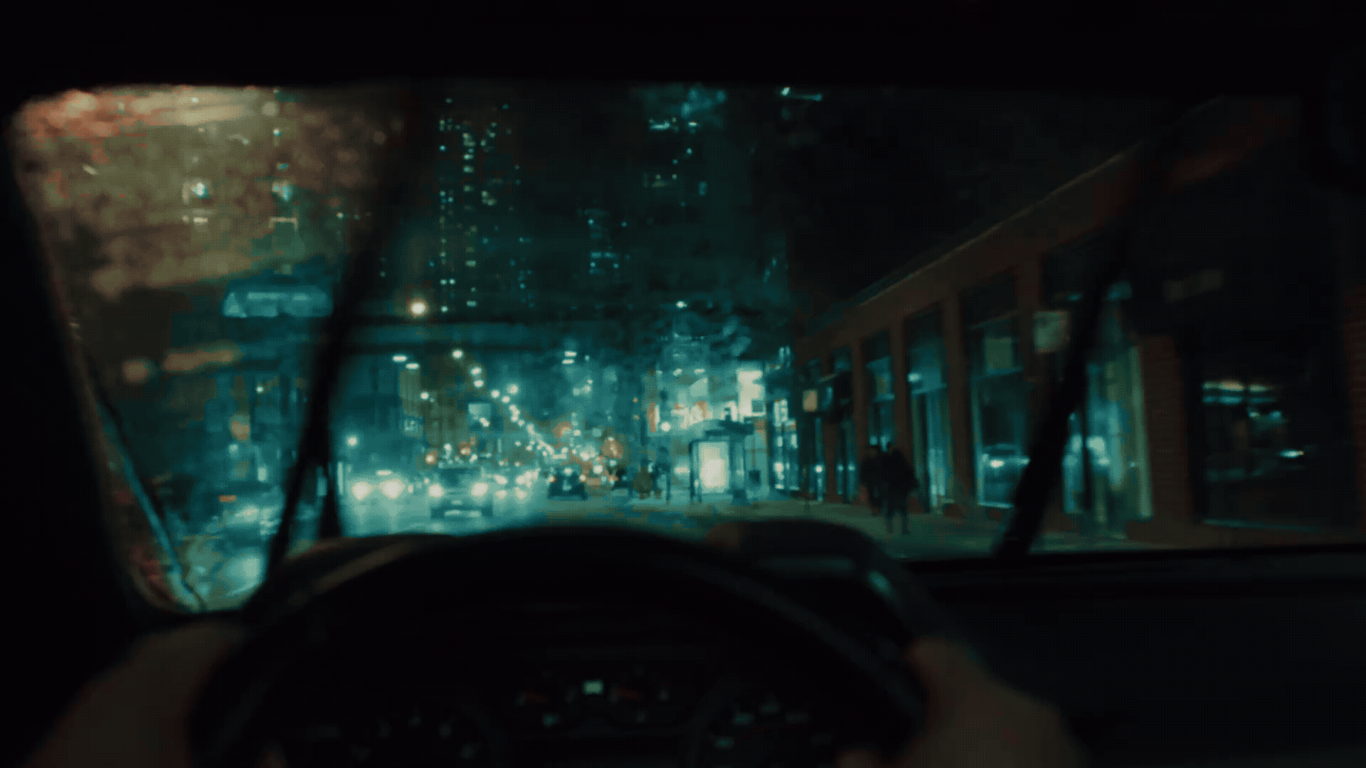 Nighttime city street scene viewed from inside a car, with rain on the windshield and raindrops visible, illuminated by city lights and traffic.