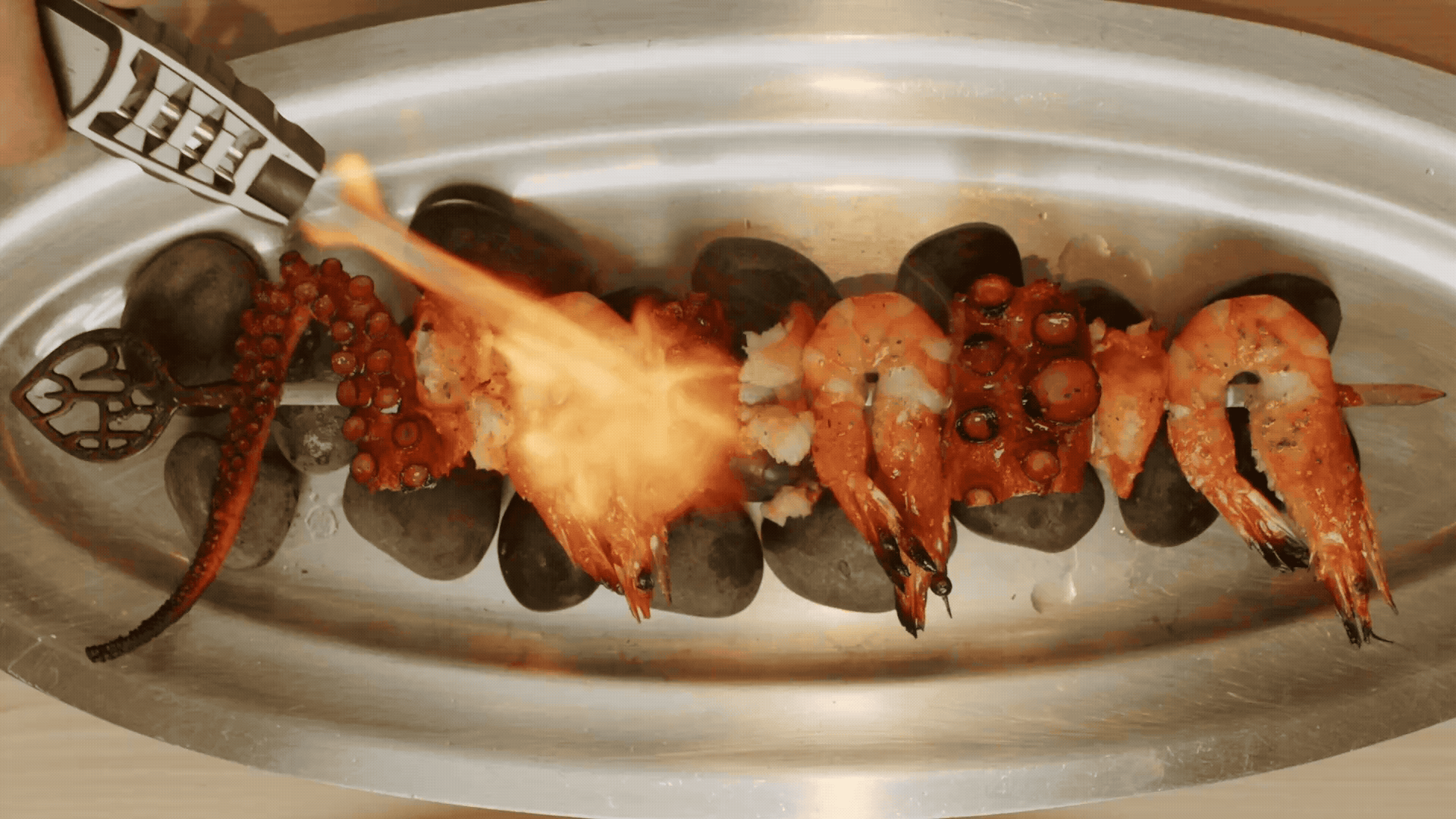 A seafood platter featuring lobster tails, shrimp, and octopus tentacles, garnished with black rocks and served in a silver dish.