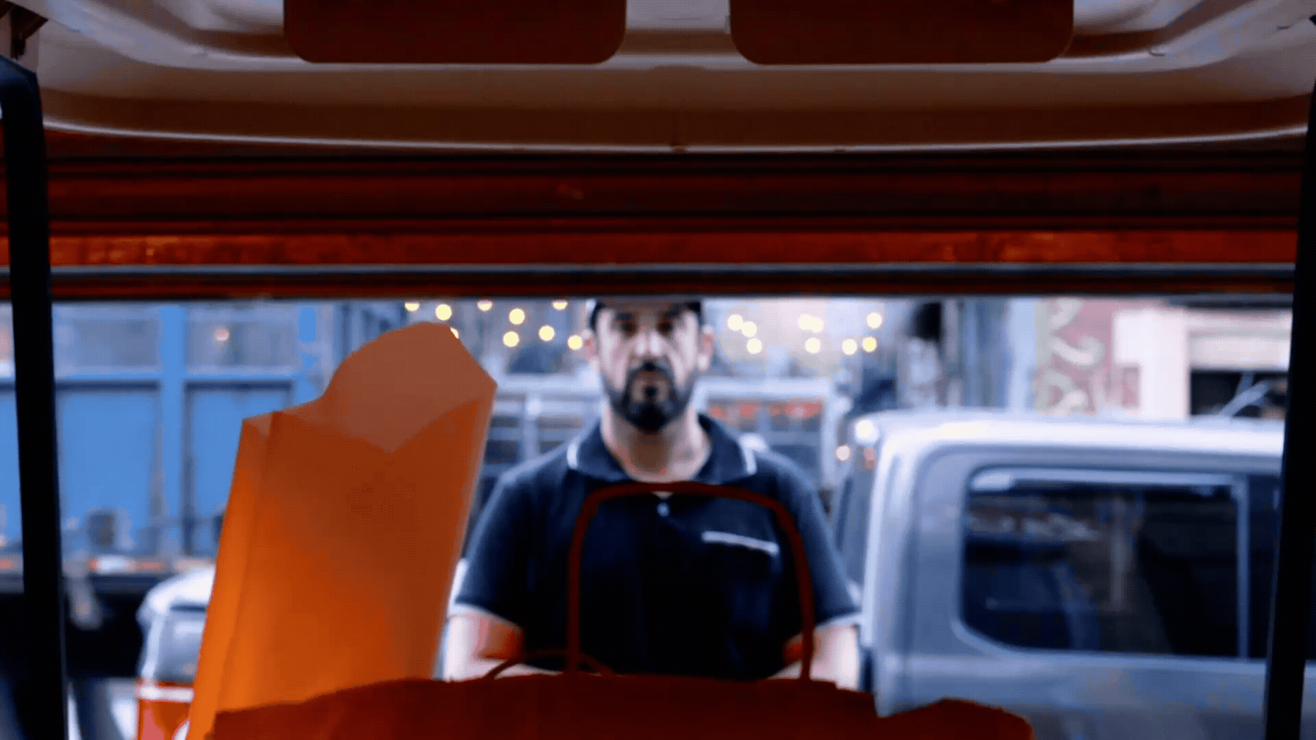 A man with a beard and dark hair looking through a service window, with an orange bag in the foreground, parked cars, and a city street in the background.