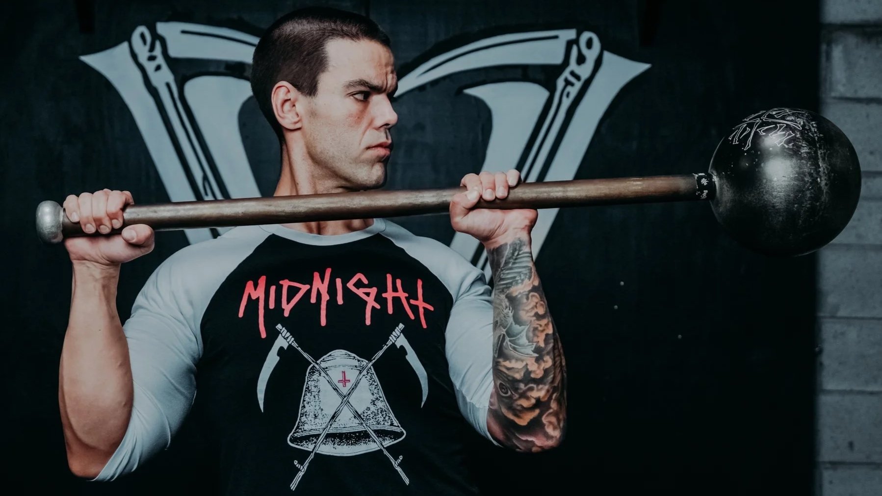 A man with tattoos on his arm lifts a Steel Mace over his shoulder during a workout in a gym.