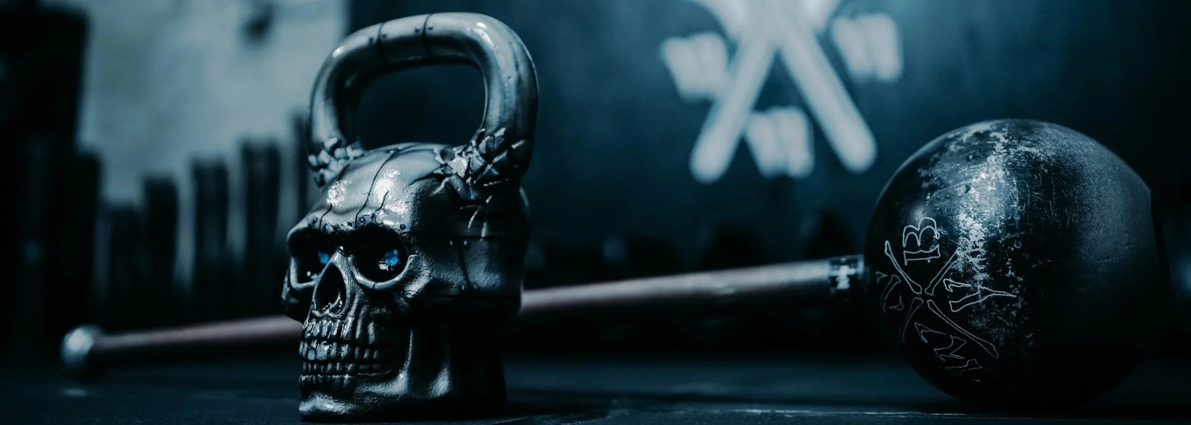 A black skull-shaped kettlebell with blue eyes, a worn black medicine ball with a skull and crossbones graffiti, and a barbell in the background on a gym floor.