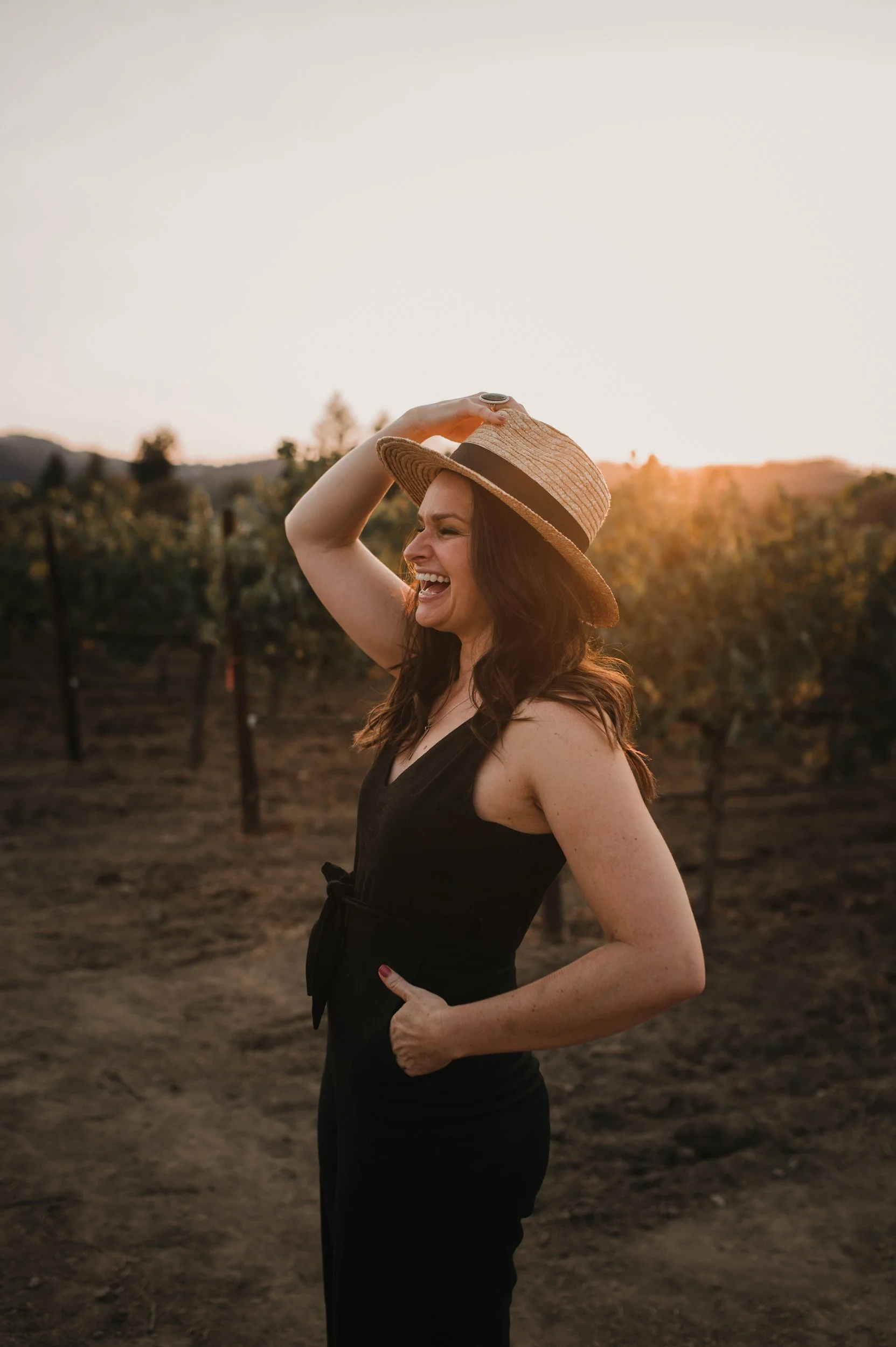 A young woman stands in a vineyard laughing as the sun sets in the distance.