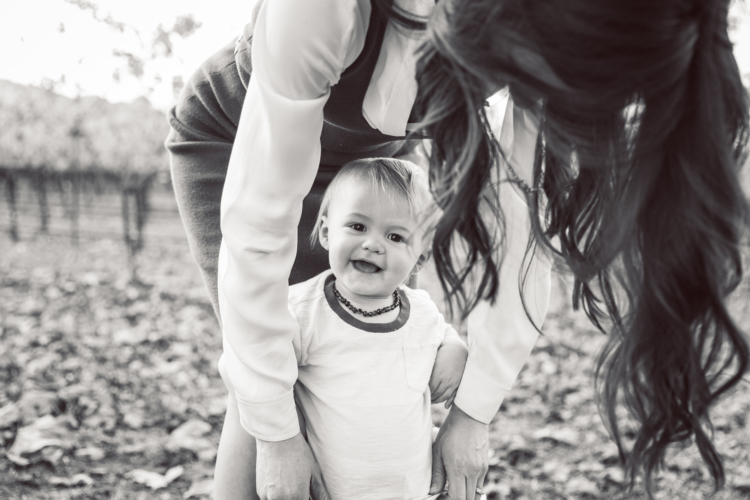 A smiling baby is standing between his mom's legs, while she looks down at his, her curly hair cascading down around him. The image is in black and white.
