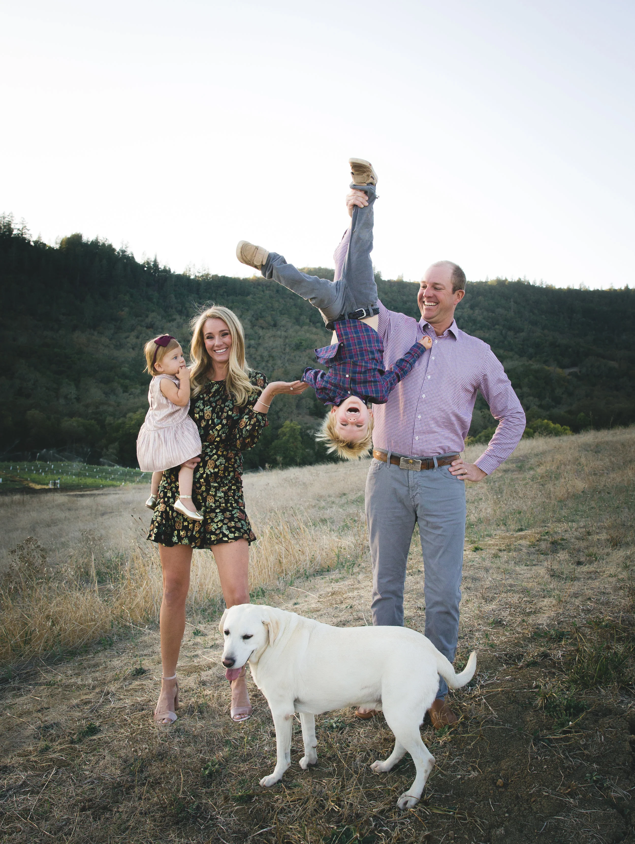  A family of four and their dog, dad is holding his sun upside down by one of his legs. Son is laughing, Mom has a funny look on her face reacting to the shenanigans. Their dog, a yellow lab is in the photo as well.