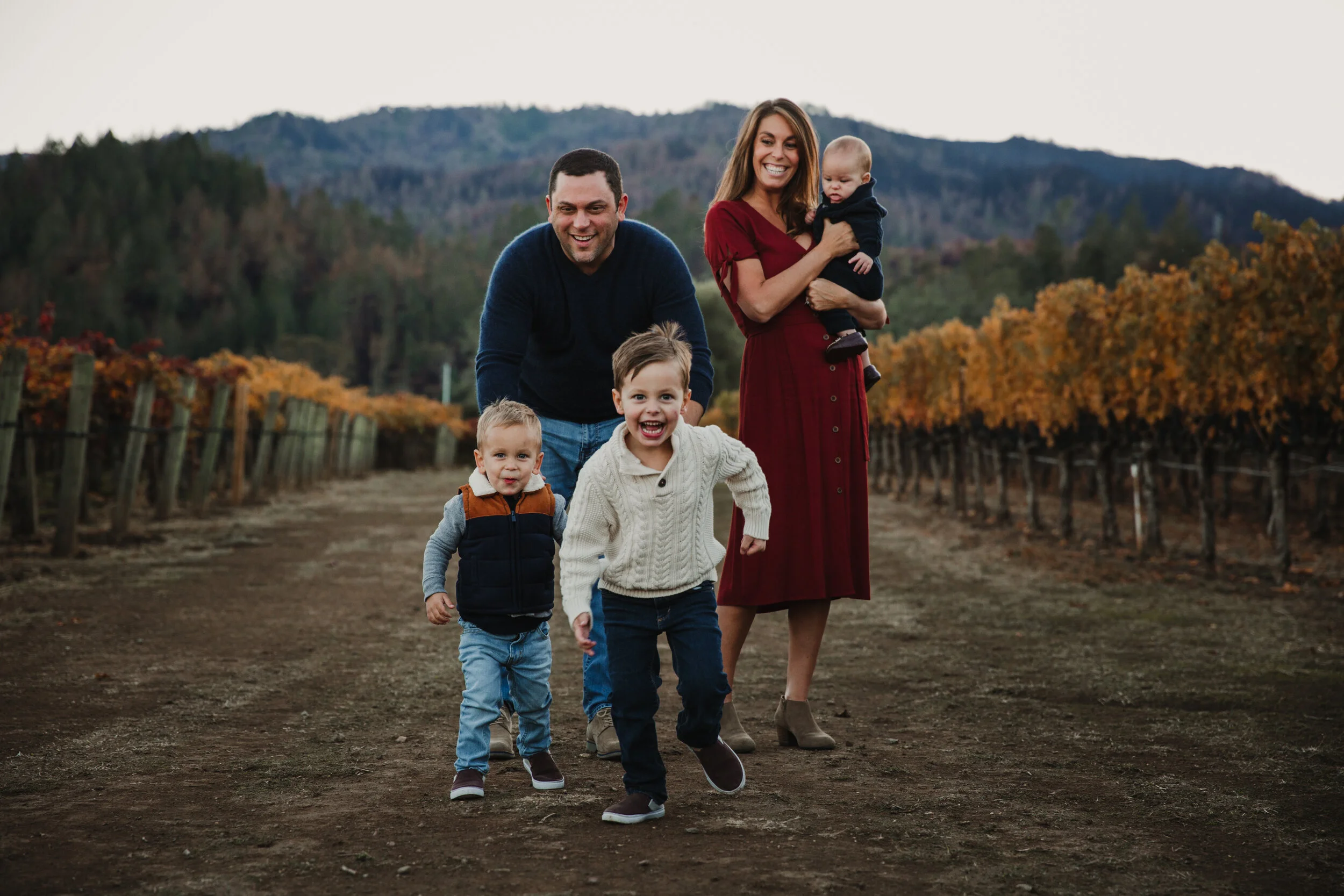 Two little boys running towards the camera, while their mom and dad and baby brother stand behind them watching and smiling. Everyone is dresses in fall colors, with orange and yellow vineyard leaves in the background. 