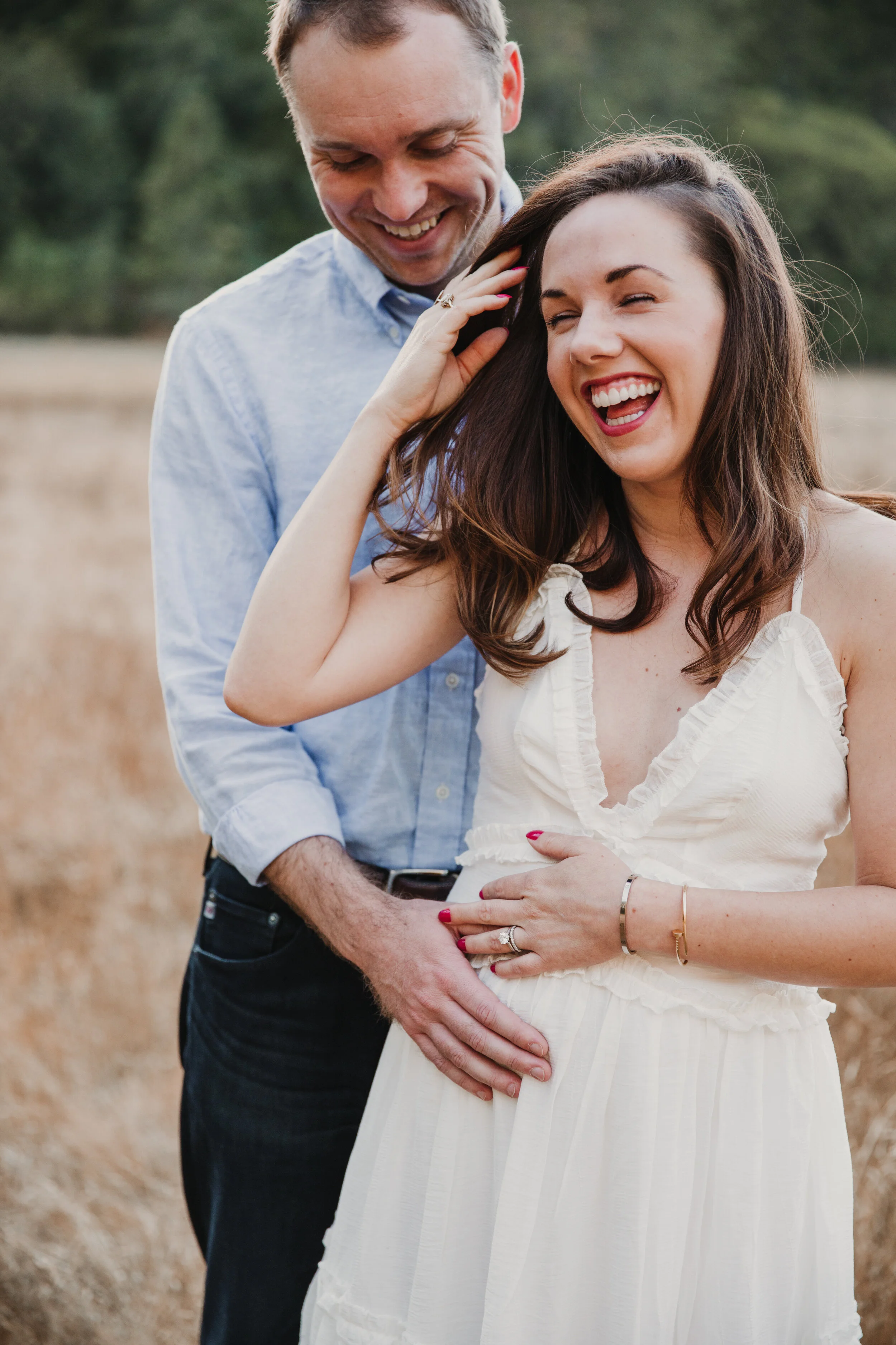 A young pregnant woman laughs and brushes the hair out of her face with one hand, the other hand on her belly. Her husband standing behind her, smiling down at her.