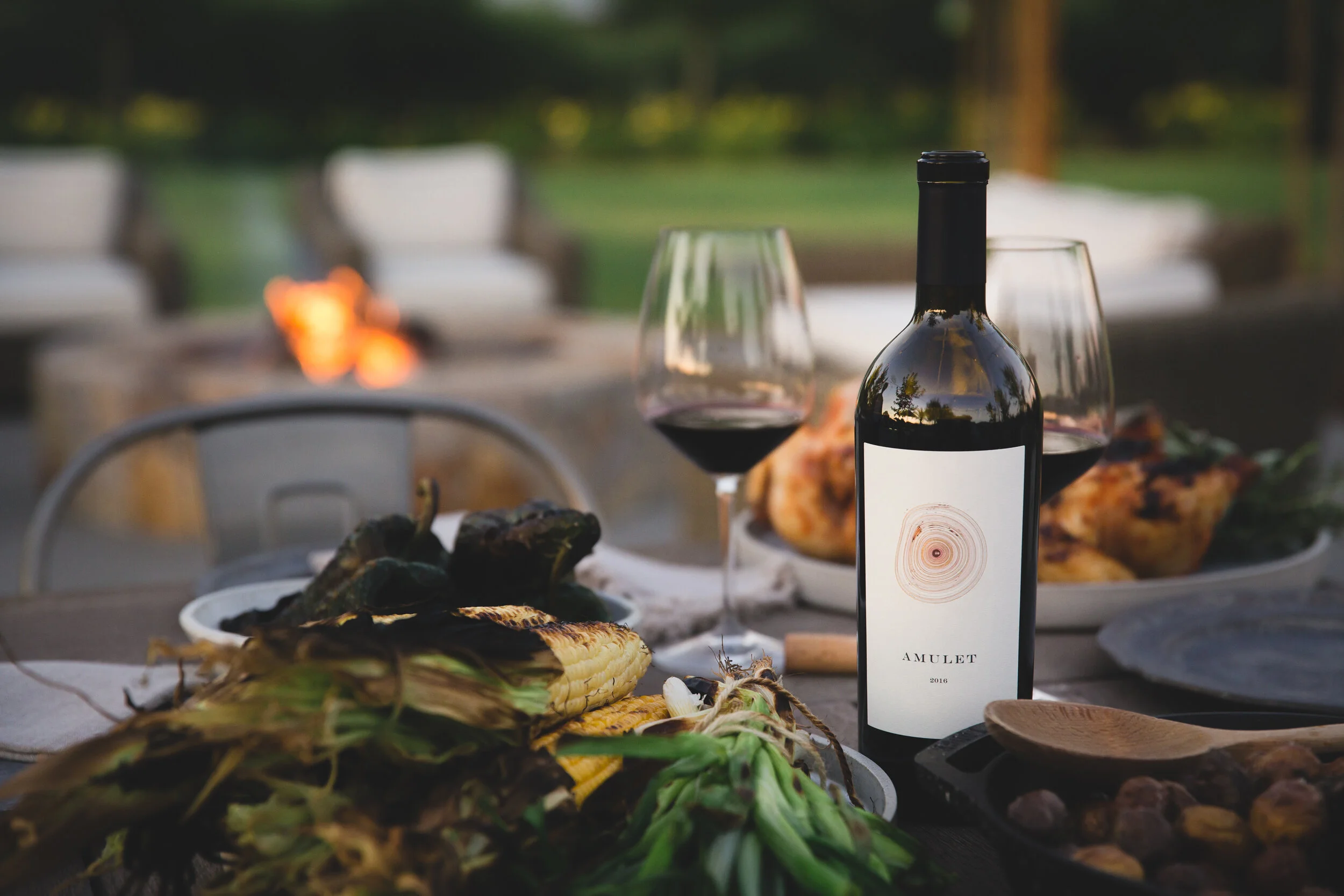 A summer outdoor table is set, and filled with barbecue, complete with a bottle of red wine. A fire pit glows in the background. 