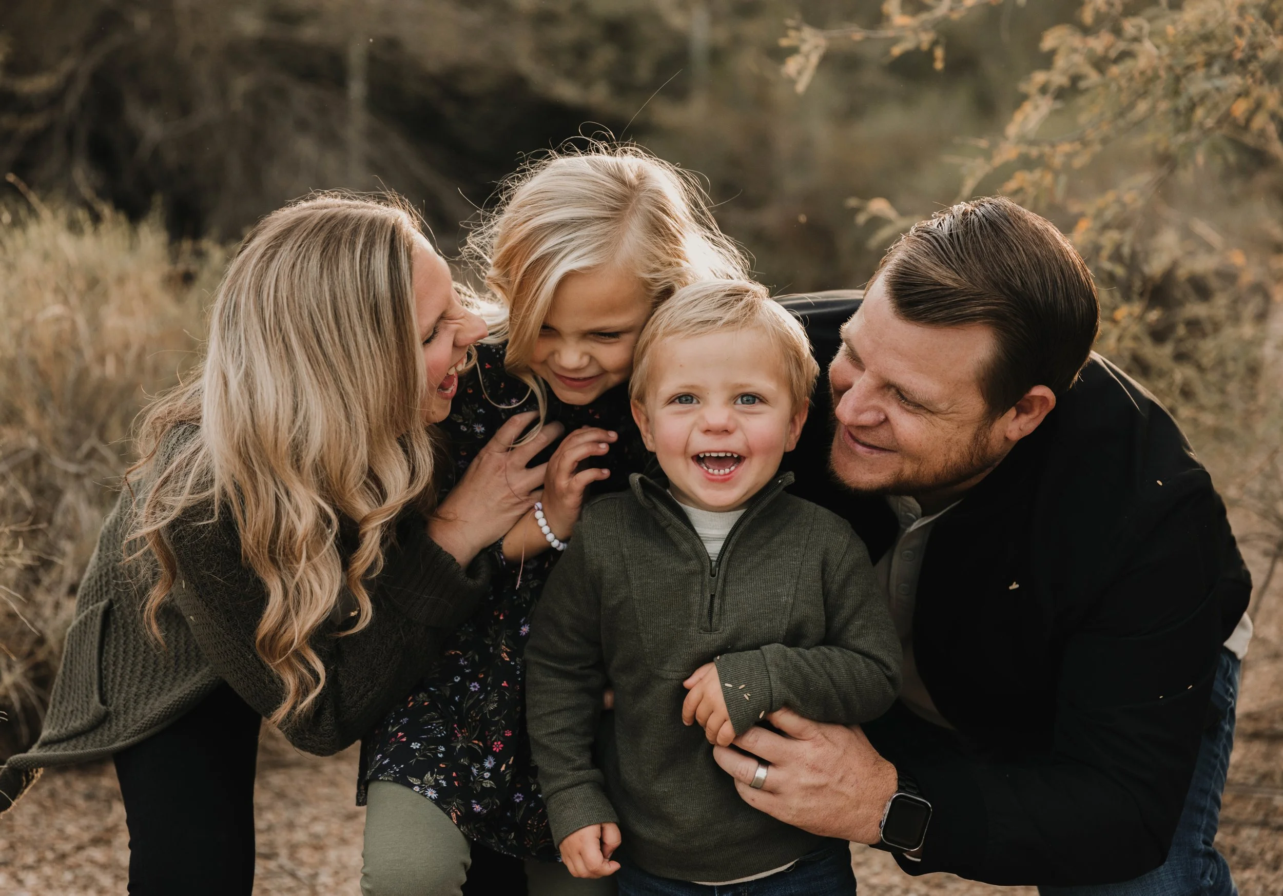 A smiling brother and sister are being hugged and cuddled by their mom and dad, golden light all around them. 