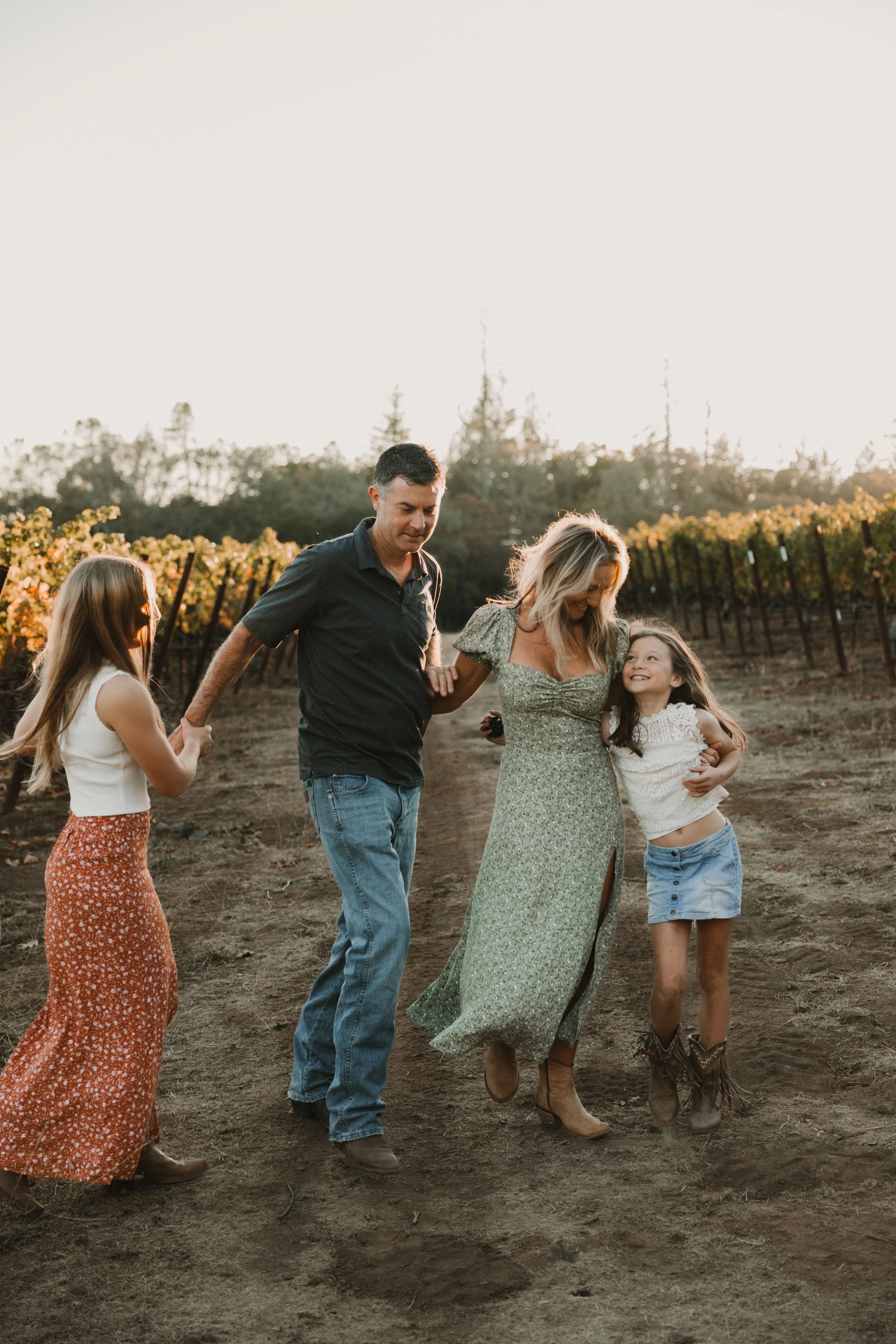 A family of four walking together in a vineyard during the fall season. There is a mom and dad, and two sisters, taken during the fall season, with yellow and orange leaves in the background. 