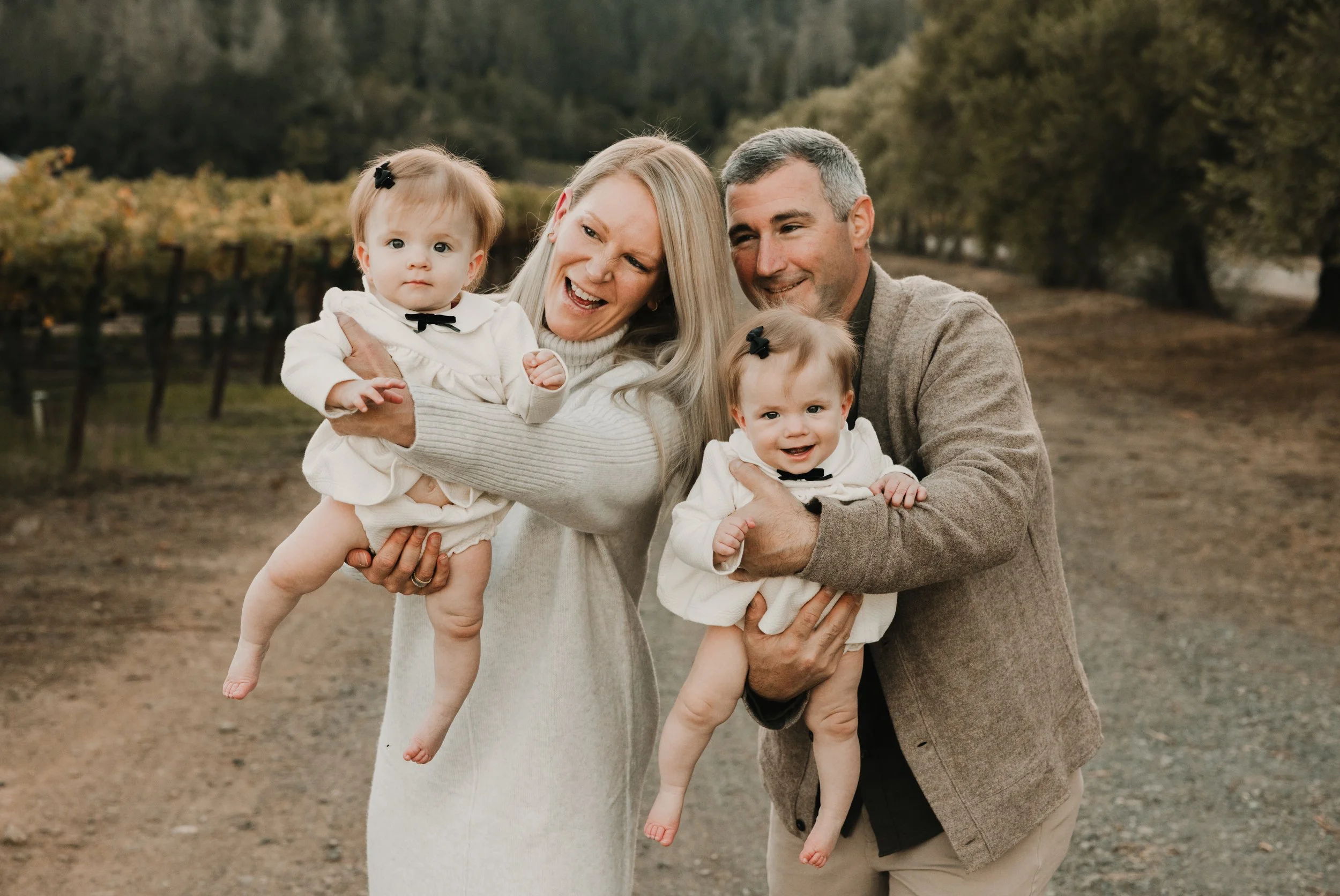 A man and woman are each holding a baby twin girl in their arms in a vineyard during the fall season. They are moving them out towards the camera. One twin is smiling, the other is looking at the camera with curiosity. 