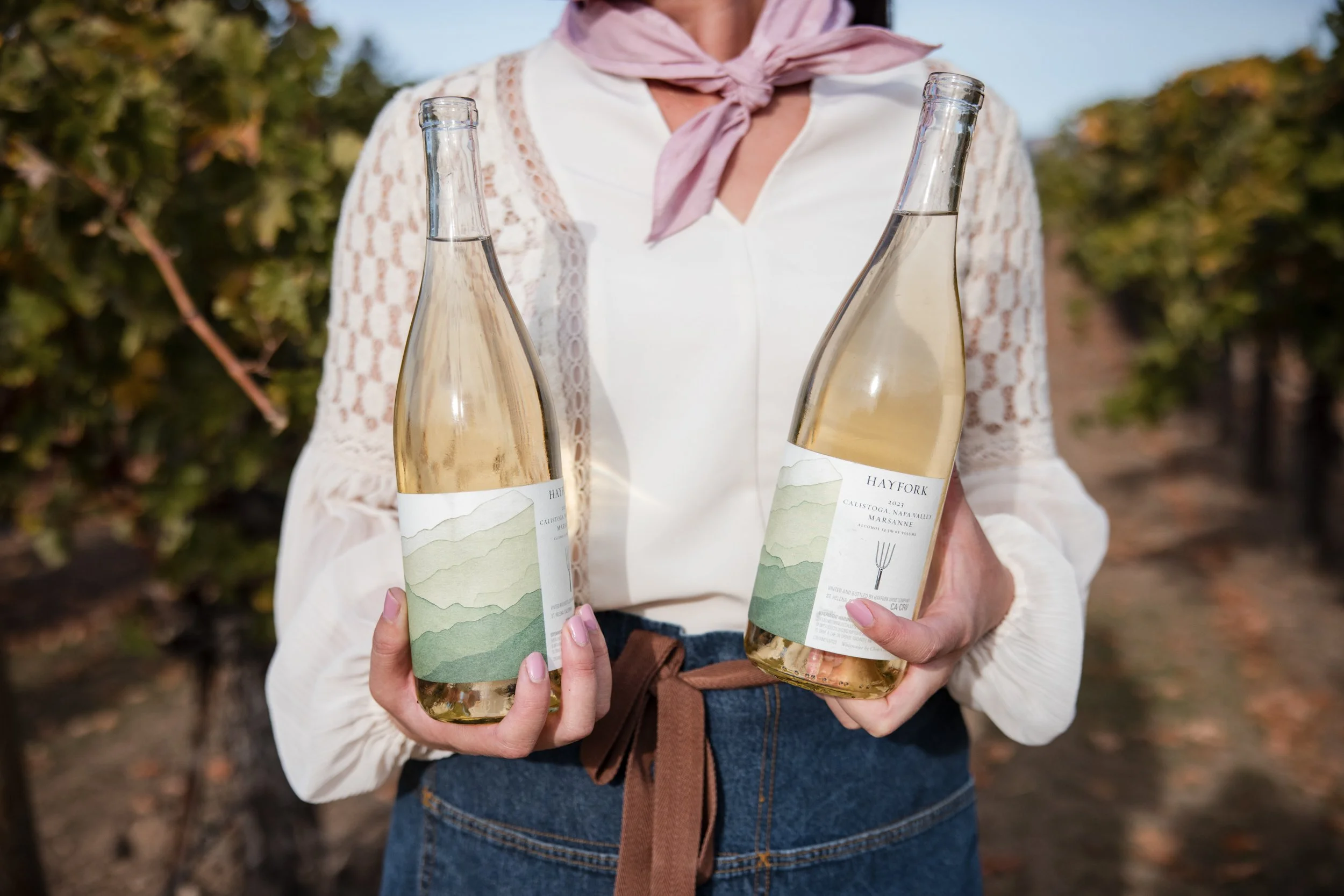 Two bottles of white wine are being held by a server standing in the vineyard. 