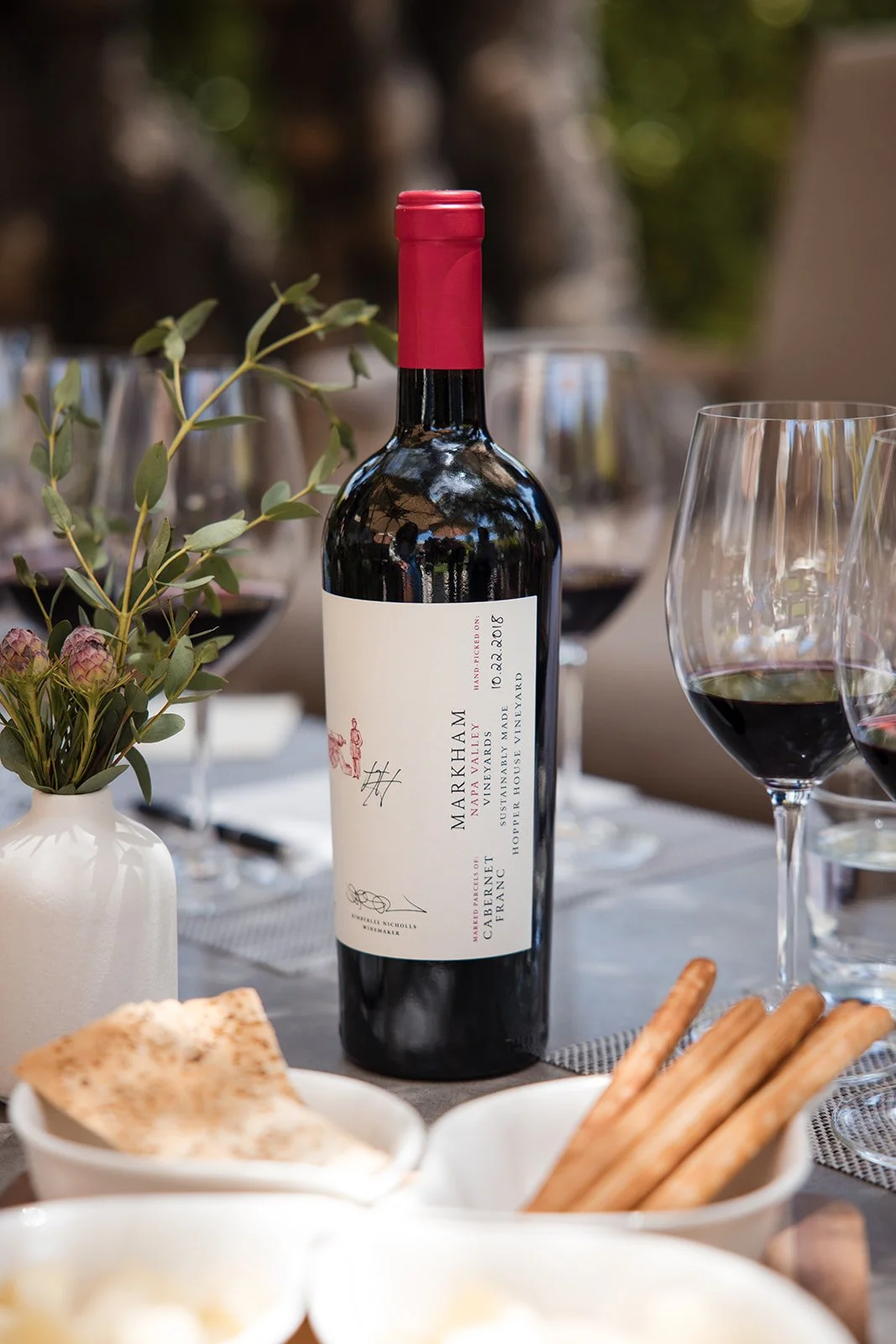 A bottle of wine is sitting on a table with a red foil around the neck of the bottle. Charcuterie and other place settings are gathered around it.