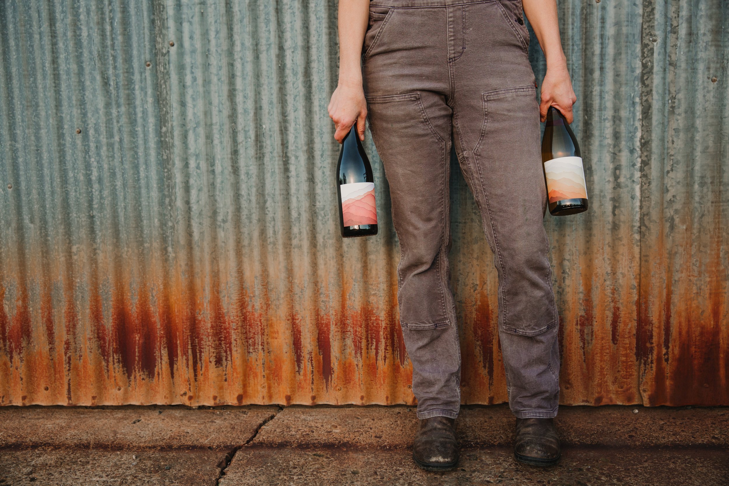 A woman holds two wine bottles against a rusted barn. 