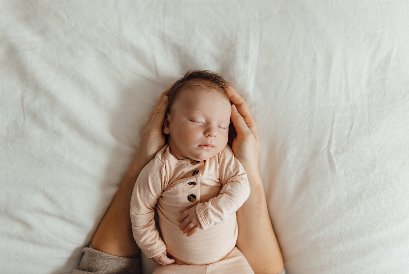 Newborn baby girl sleeping on a bed, her mom's hands cradling her head. She is wearing a cute pink onesie with brown buttons.