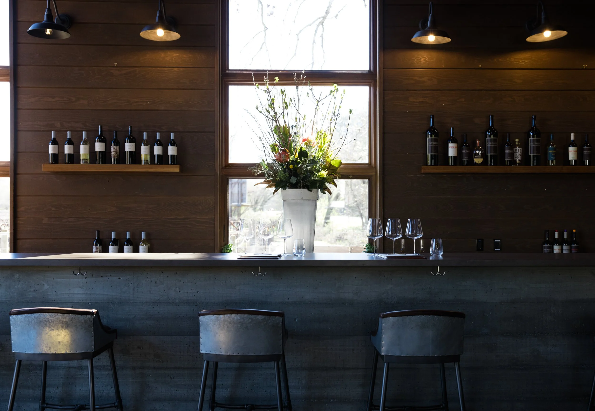 A high end Napa tasting room is set up, ready for visitors. Wine bottles are lined up on the walls, and the bar stools are open and table settings set. 