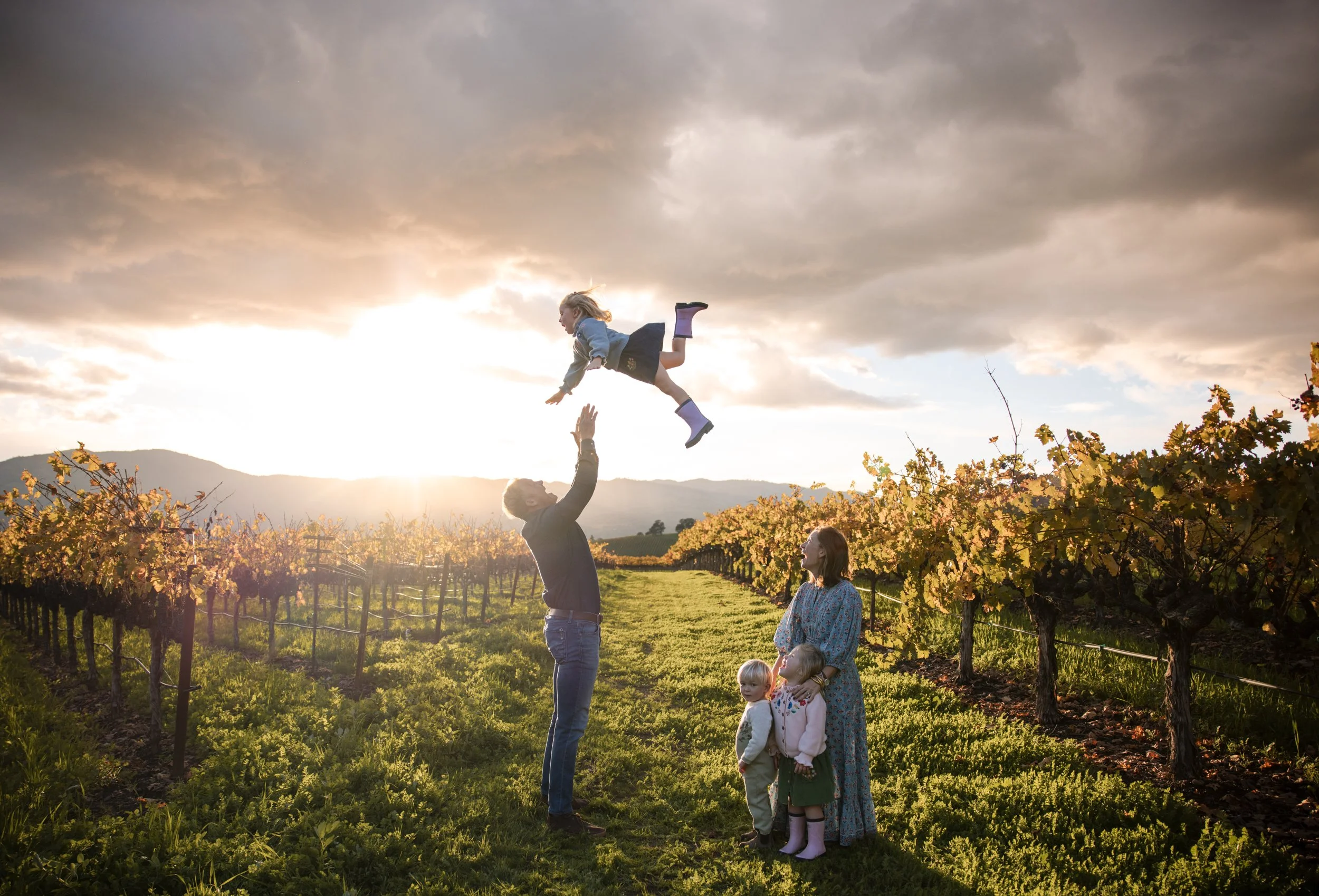 A little girl wearing rainbows is being thrown into the air by her dad in the vineyard as the sun sets behind them. Mom, little brother and sister are standing nearby smiling and watching.
