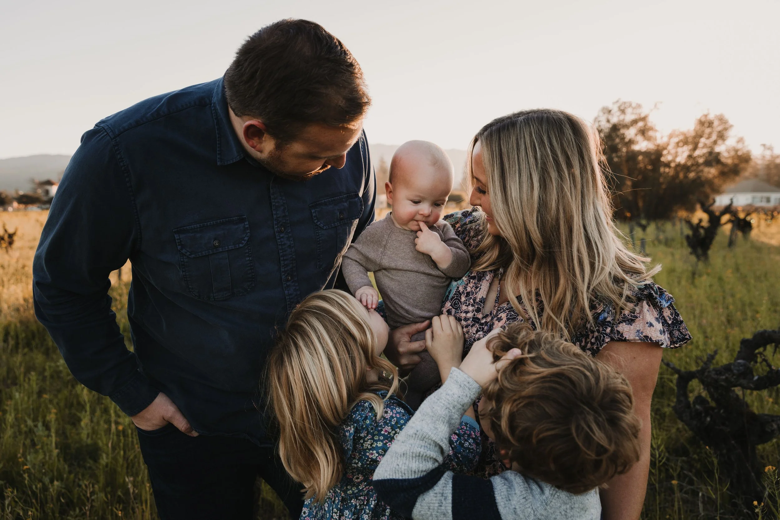 A mom is holding her baby next to her husband and two other children in a green field. Everyone is looking at the cute baby. 