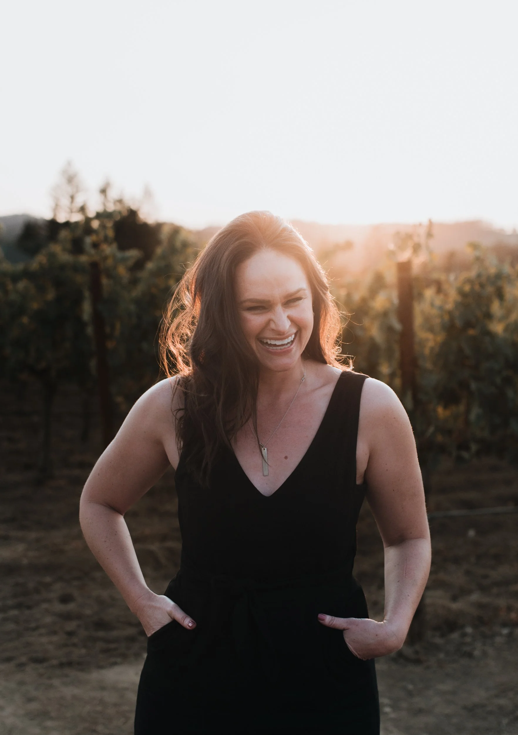 A young woman in the vineyard smiling, while the sun sets behind her.