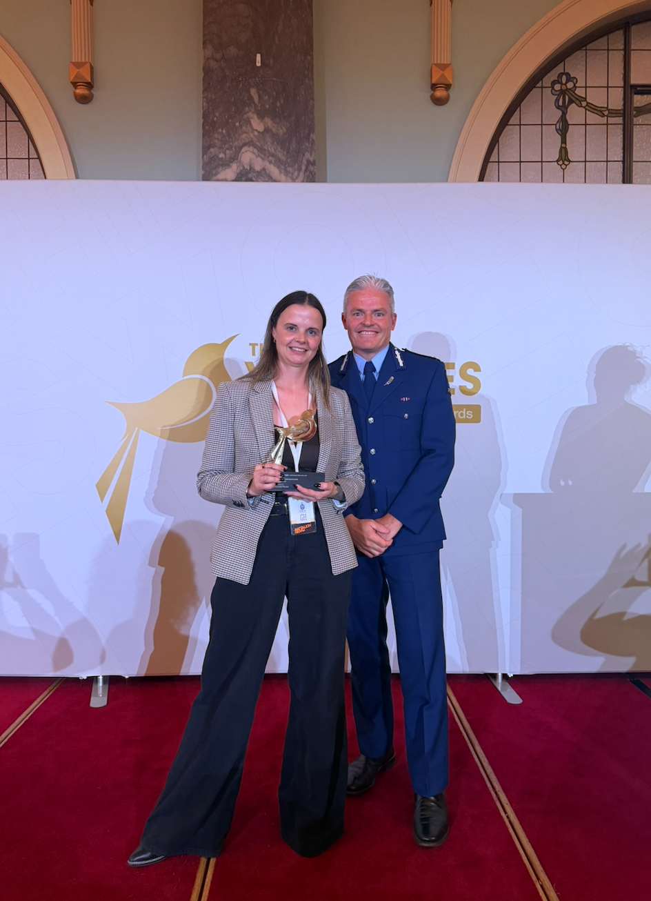 A woman holding an award standing next to a man in a blue military uniform at an award ceremony, with a white backdrop featuring a gold logo and text.